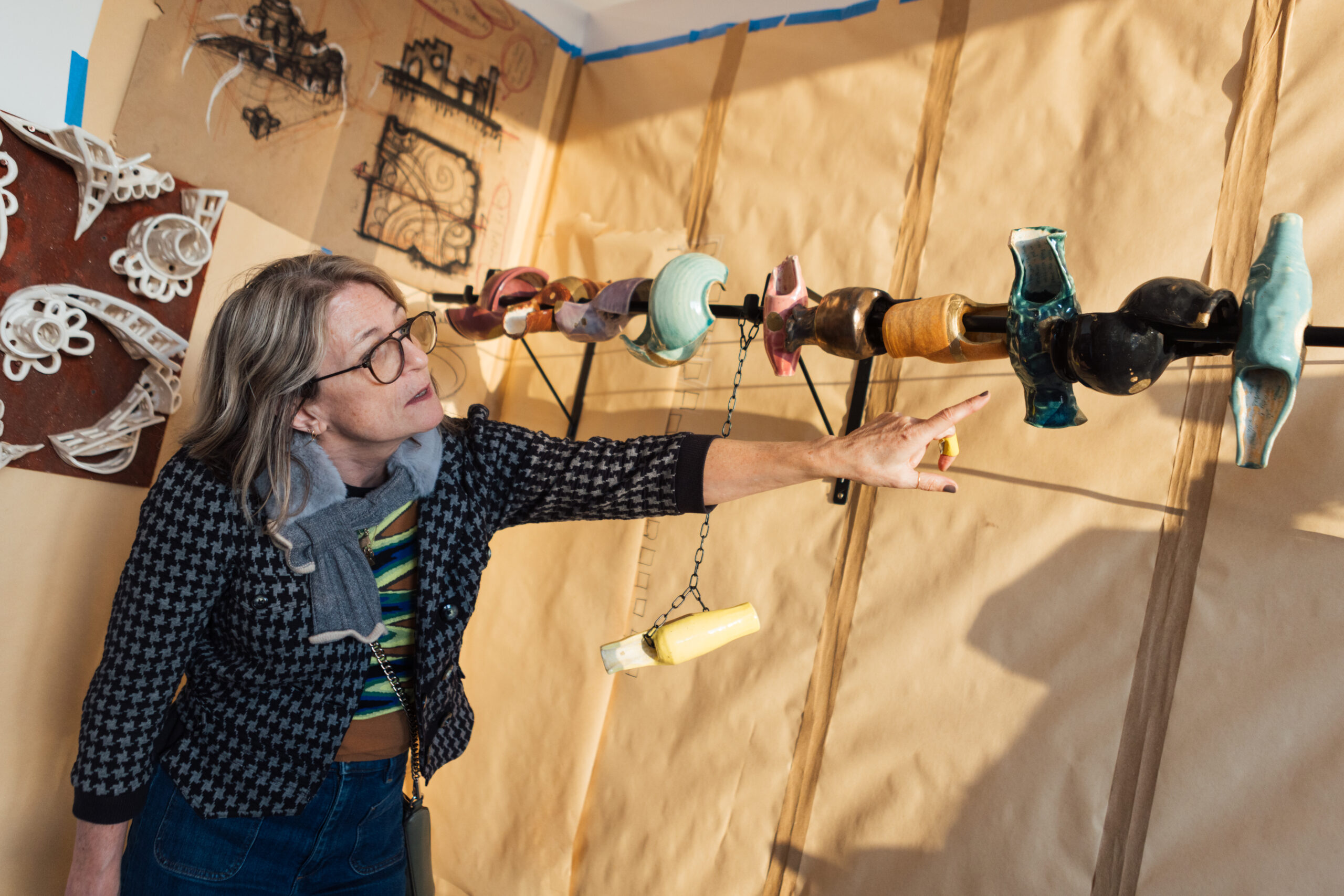 A woman wearing glasses, a scarf, and a blazer points to an artwork installed wall-to-wall across an MFA open studio. The walls are all covered in beige. The artwork is sculptural objects on a rod.