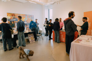 A group of people in a gallery bay at Dock 72. The left table is covered in a black tablecloth and a large, colorful banner hangs on the wall behind it. The table to the right of it is covered in an orange tablecloth and an orange vertical banner stands next to it. The cropped table toward the right foreground is draped in a light purple tablecloth. All tables are at least partially obscured by standing visitors.