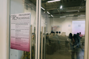 Closed double doors, entrance to the Dock 72 Fine Arts Performance Lab. On the left door is a light purple to medium purple poster with a program schedule for Open Fields. Through the other door, seated audience members can be seen.