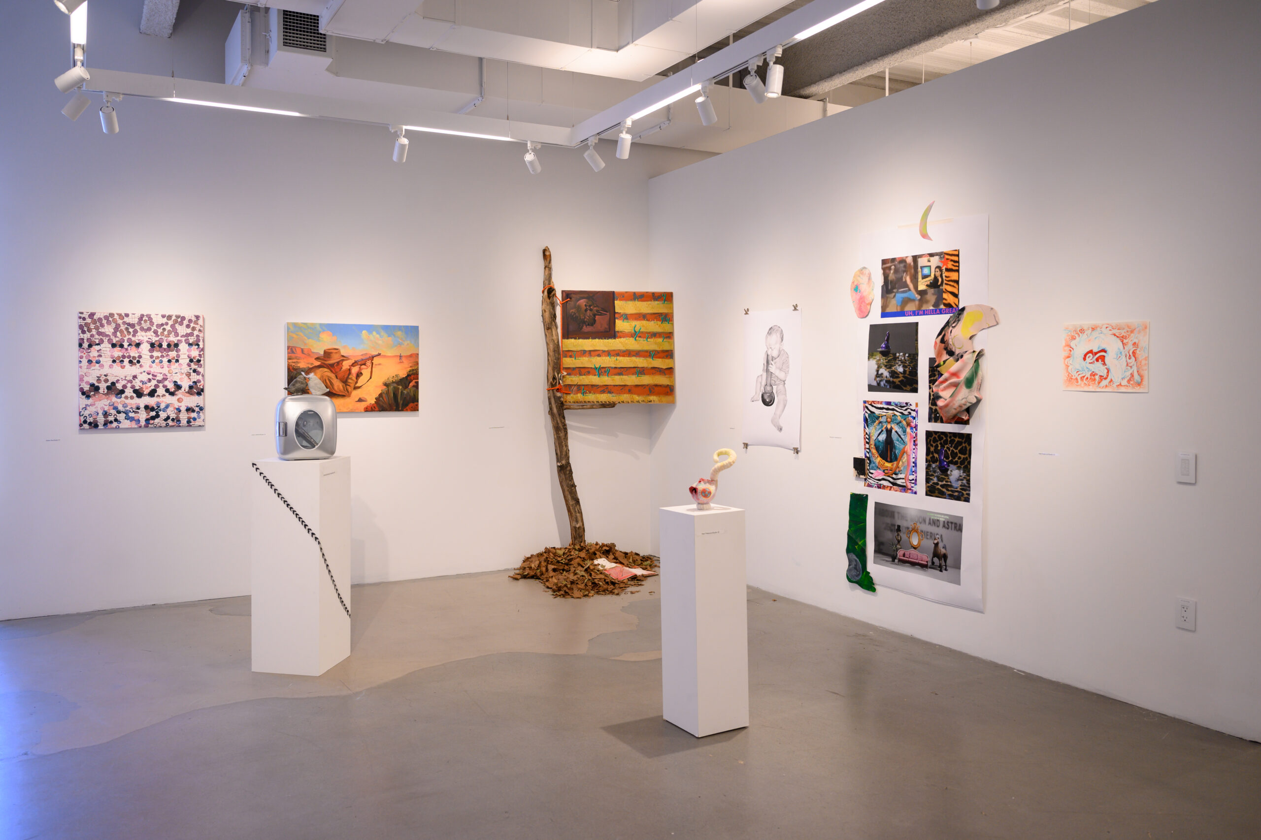 A brightly lit gallery with sculptures on pedestals. On the back wall are two 2D works. In the back corner is an American flag leaning against the wall with a pile of soil at the base. On the right wall is a group of stacked works between two smaller drawings.