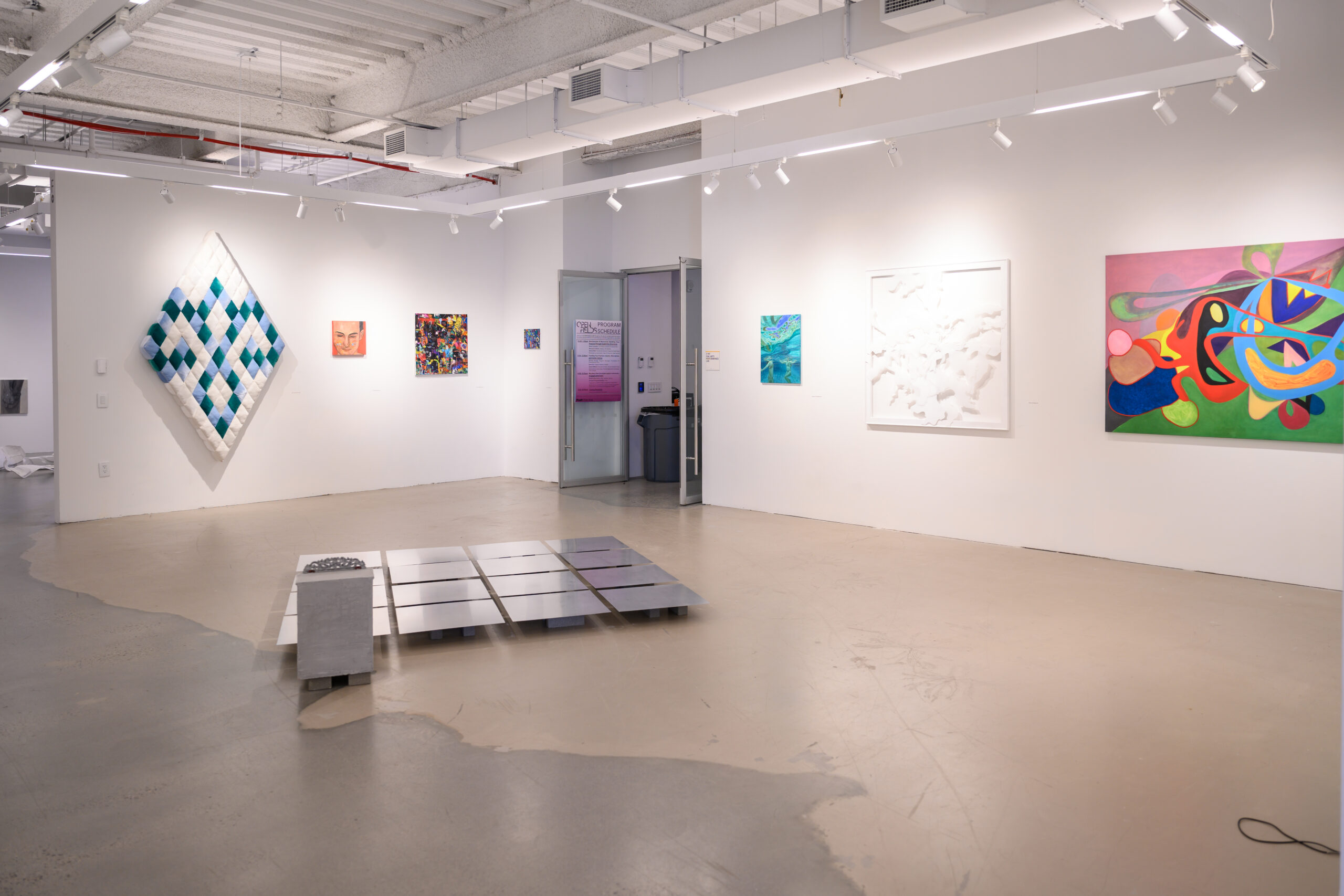 Installation of the 2025 MFA Pop-Up Show at Dock 72. A brightly lit gallery. On the back wall is a large, diamond-shaped artwork which looks quilted in white and blues. Next to this work are smaller abstract paintings. The door to the Performance lab is open, with a poster of the Open Fields panel schedule posted. On the right wall are a small blue painting, white artwork, and colorful abstracted painting. In the center of the room, on the floor, is a grid of aluminum plates.