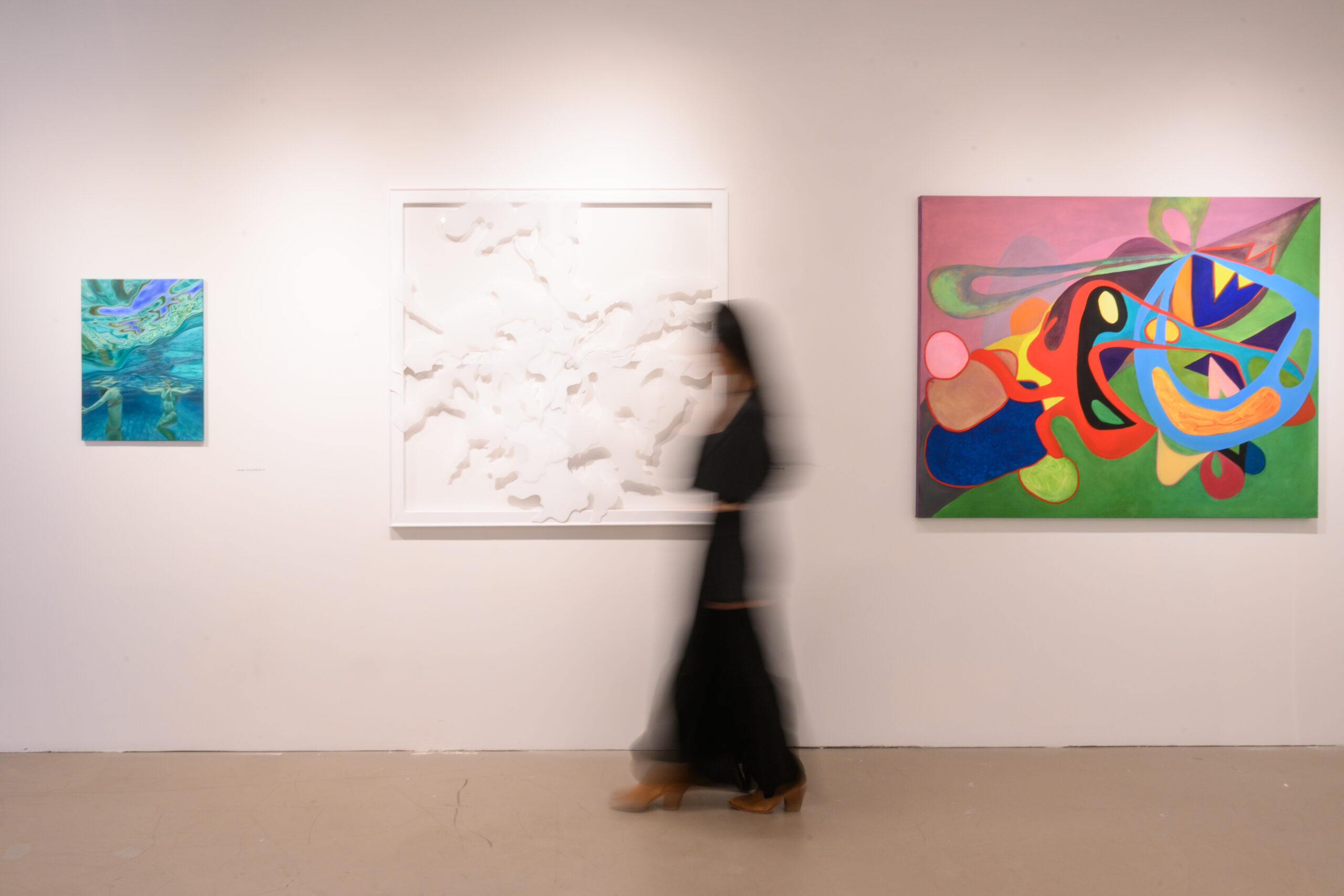 A blurred figure wearing all black walks toward the left of the frame in front of artwork hanging on the walls. The works left to right are a small blue-colored panel, an all-white square work, and a colorful abstracted painting.