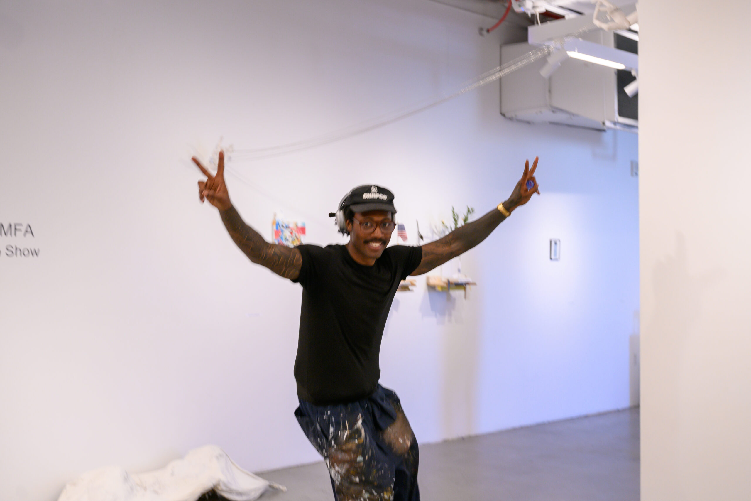 A young Black man wearing headphones, a hat, black t-shirt, and paint-covered paints, holds up two peace signs.