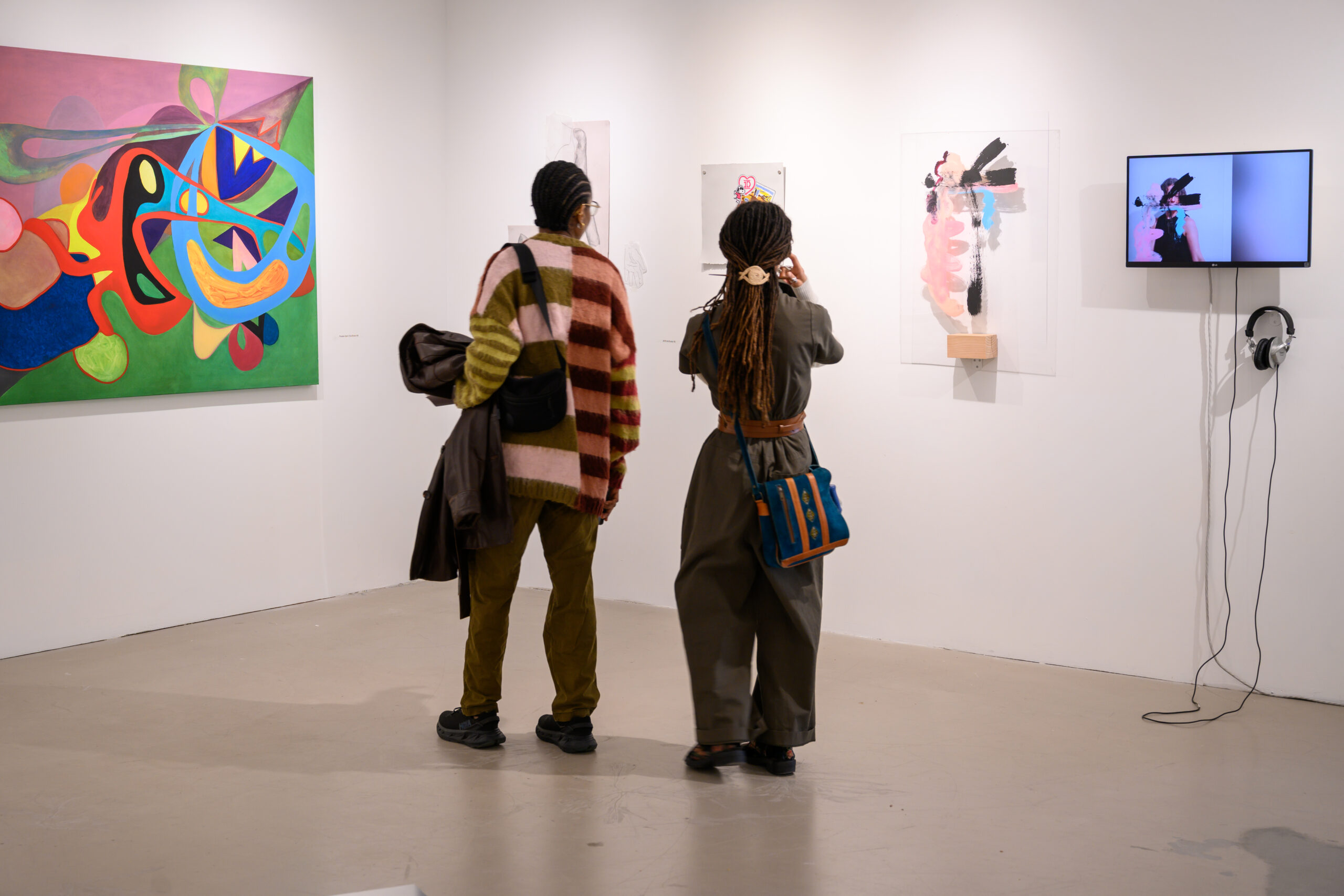 Two visitors view the 2025 MFA Pop-Up Show. The painting to the far left is a colorful, abstracted work. To the right is an abstracted work on a clear sheet and a monitor playing a time-based work, with headphones.