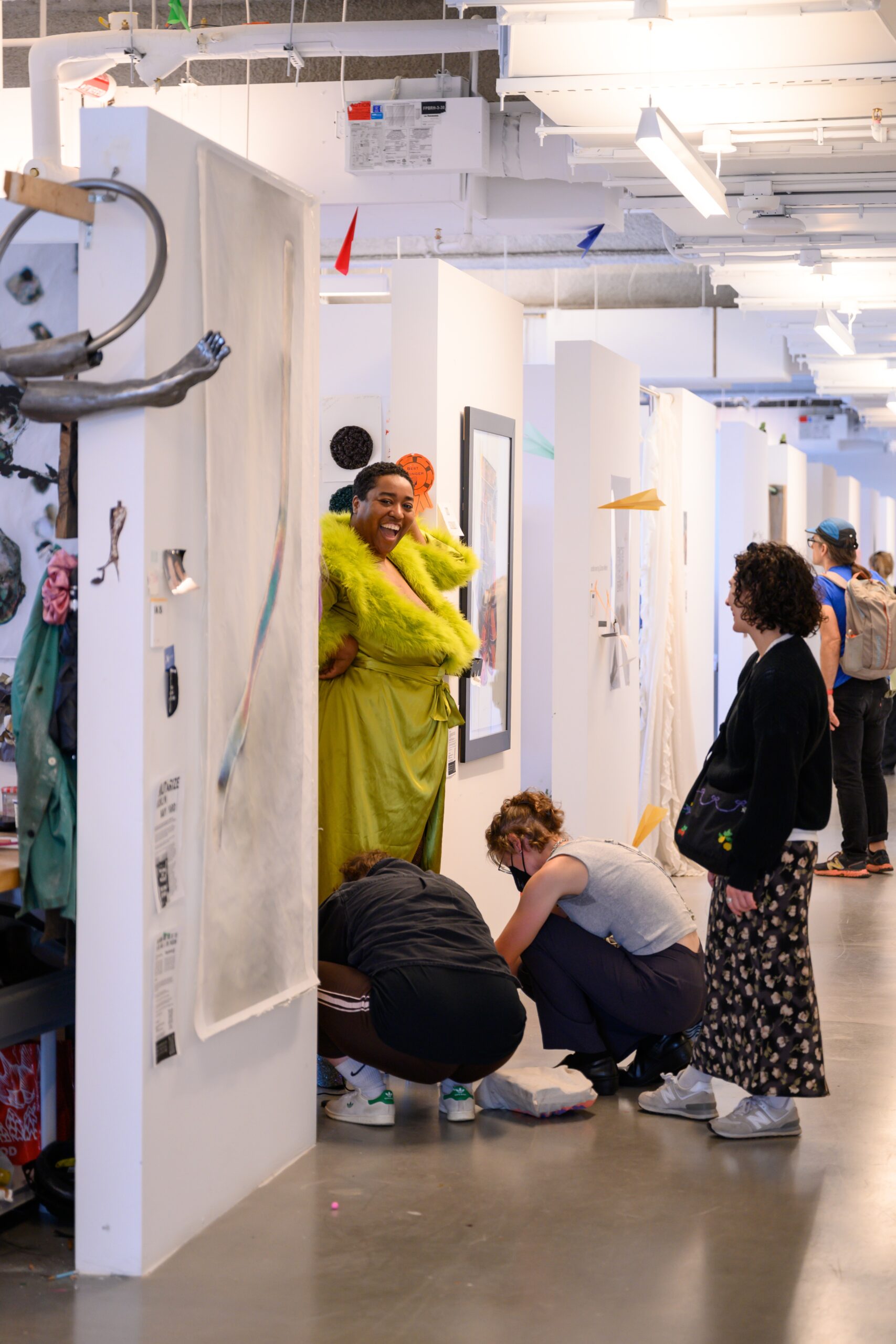 Lo Smith, wearing a green robe with fur collar, short hair, and a big smile, with two helpers at their feet assisting with arranging their dress. A visitor watches to the right of the photo frame.