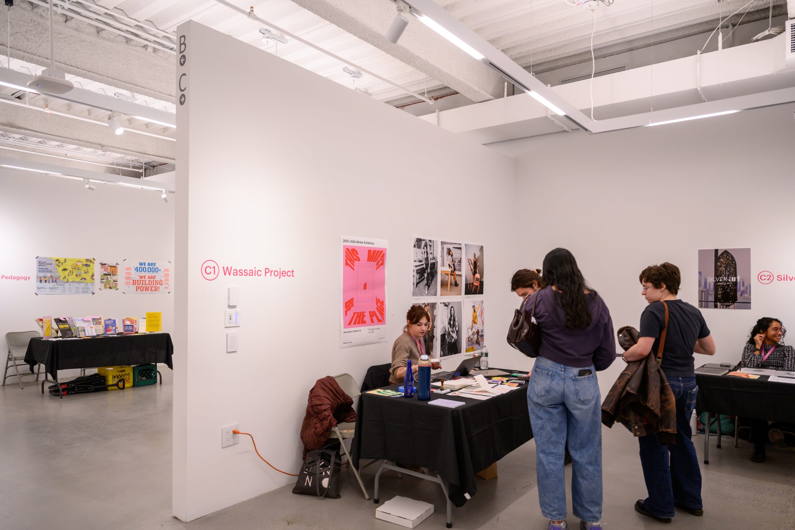 A group of people are in front of a table where a representative of Wassaic Project is seated at a black tableclothed table in front of posters on the wall. To the left, behind the wall, a table covered with brochures and pamphlets is in front of a wall with posters. Directly to the left of the visitors is another Open Fields table.