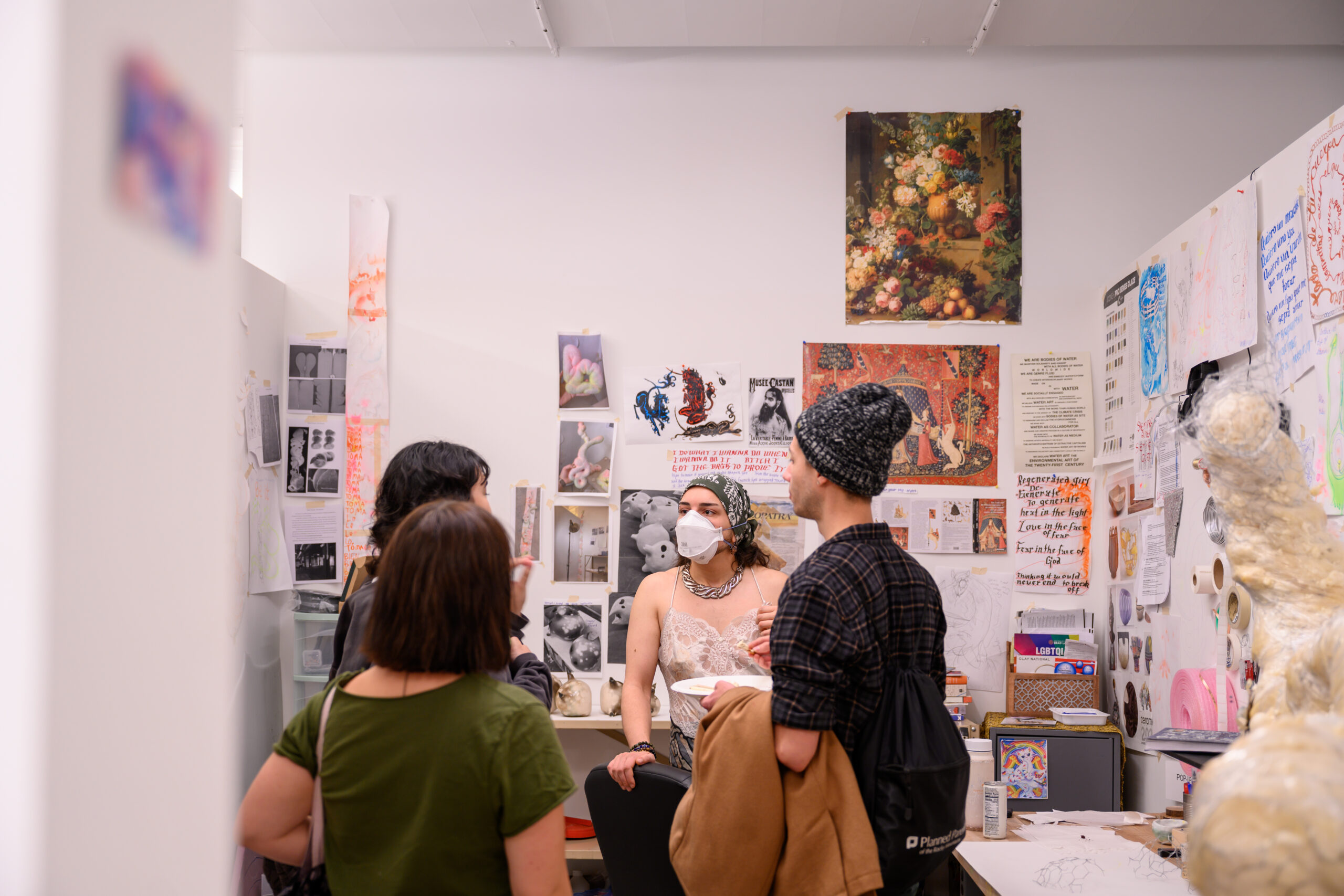 A group of visitors in an MFA studio at Dock 72, speaking to the artist, who is wearing a mask. The wall behind them has different 2D works posted on them up to the ceiling.