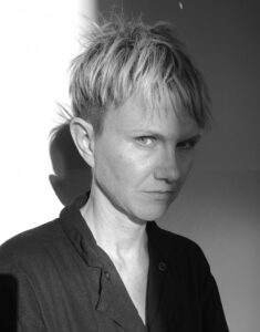 Black and grey portrait of Laurel Sparks. They are in the center of the frame and lit from the beyond the left side of the frame. Their head is tilted downward at 3/4 view, and their gaze is pointed at the camera.