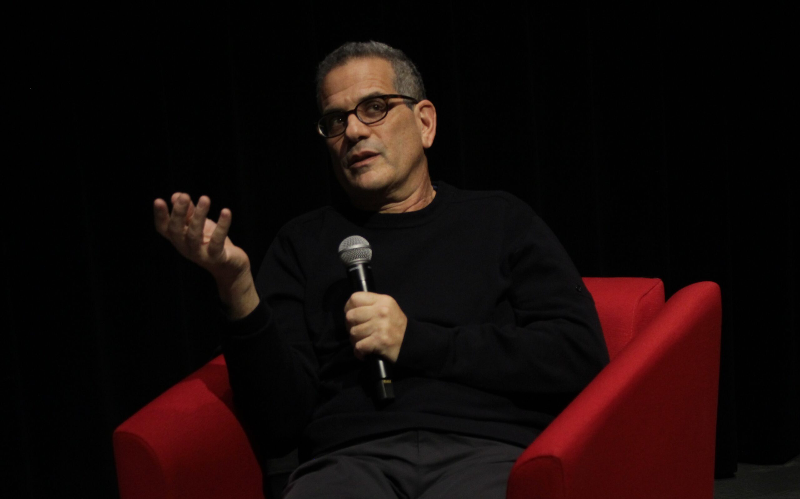 A man seated in a bright red armchair on a dark stage holds a microphone in one hand while gesturing with the other. He wears glasses and a black long-sleeve shirt, and he appears to be speaking to an audience. The background is completely black, creating strong contrast with the red chair.