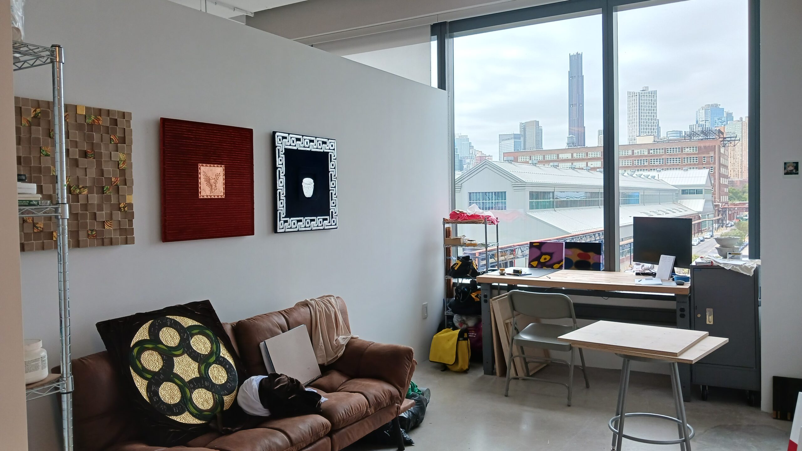 A Fine Arts MFA studio at Dock 72. Three works are installed on a wall, under which there is a couch and cushions. A table is against the window toward the back, which overlooks the Brooklyn Navy Yard.