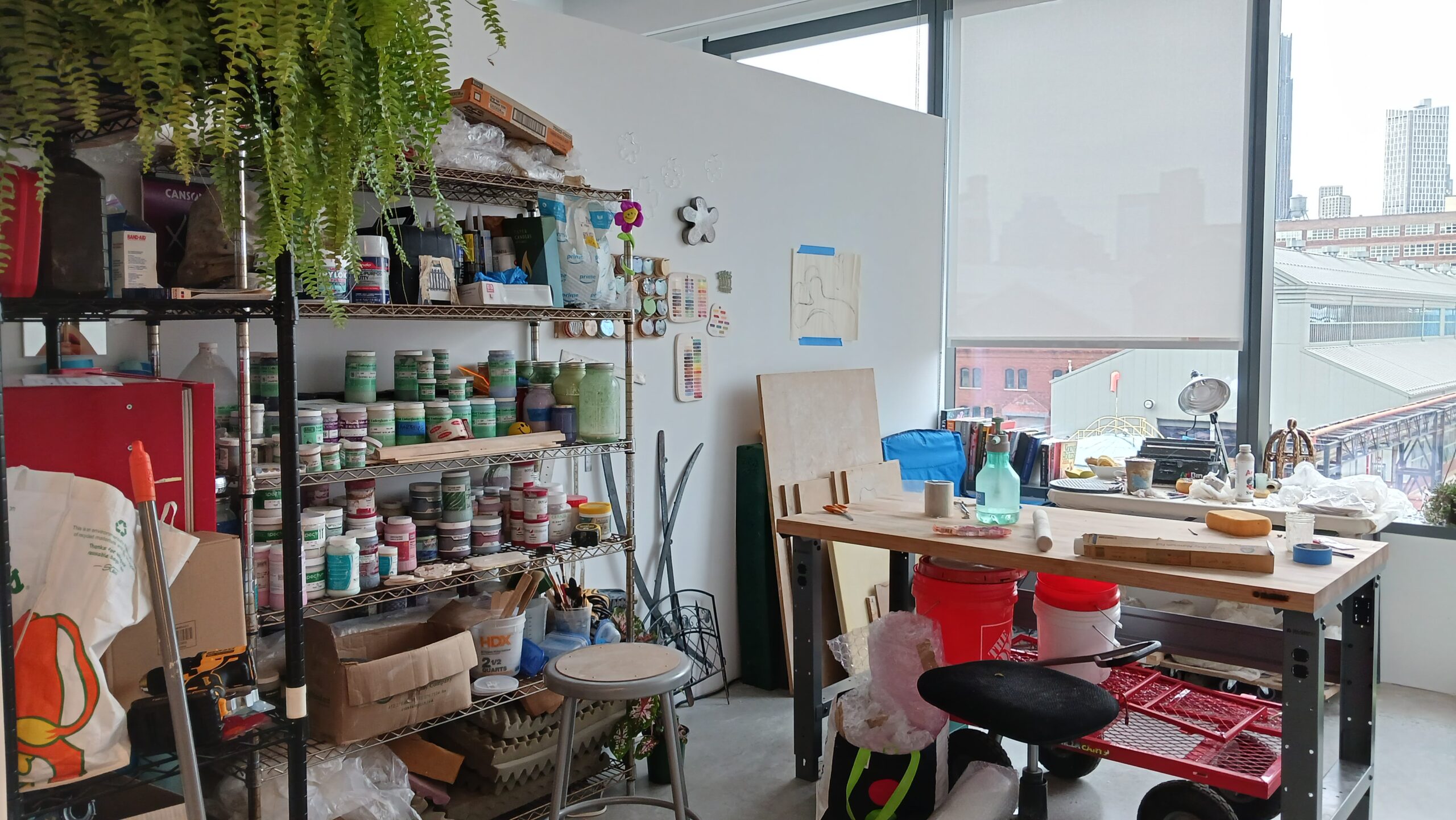 Fine Arts MFA studio at Dock 72. The left is a shelf with art supplies and large plant. In the center is a table with art supplies on it. Behind is a window overlooking the Brooklyn Navy Yard.