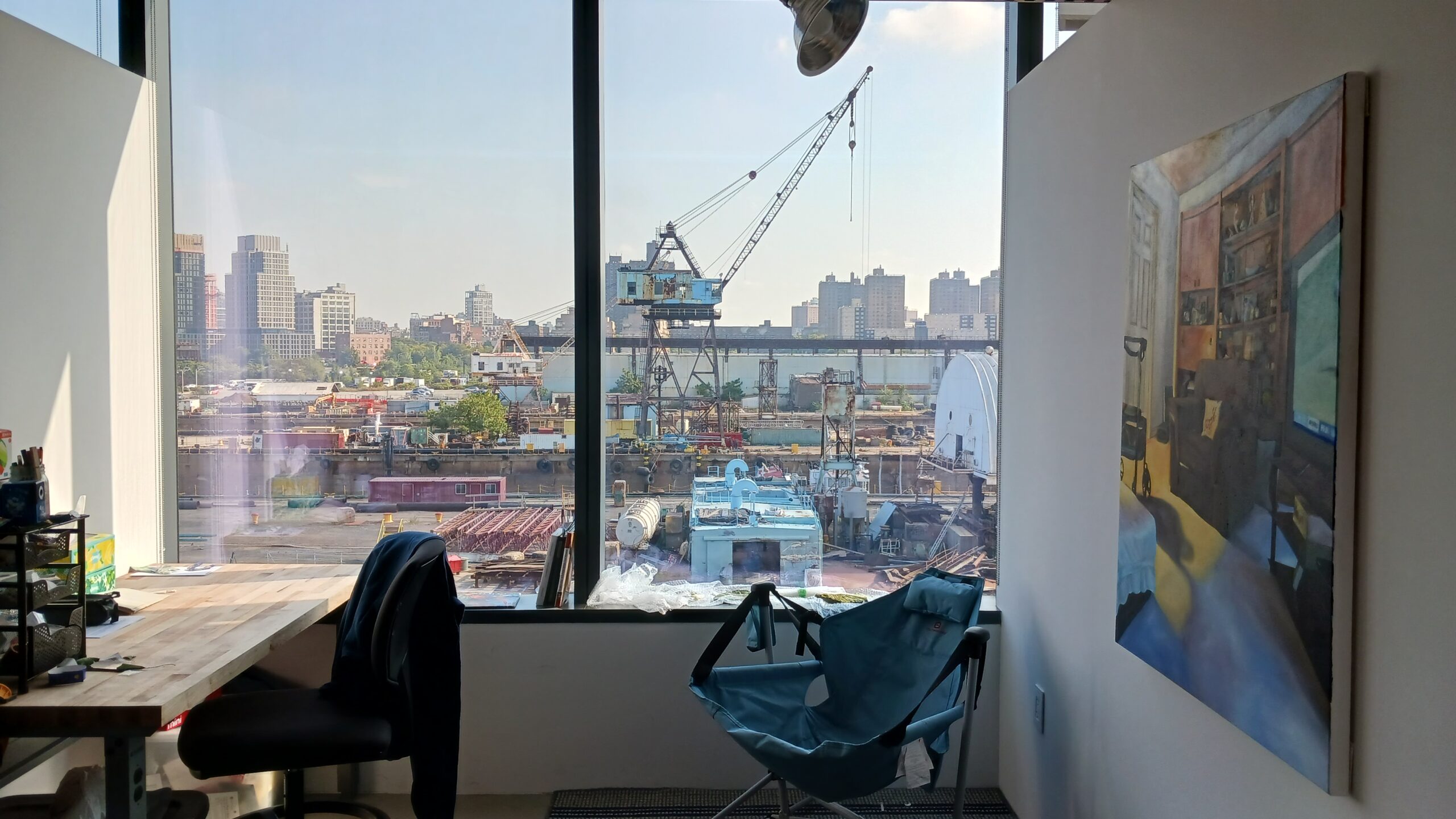 A Fine Arts MFA studio in Dock 72. A desk is positioned against the left wall with a rolling chair. There is a folding chair in the back right corner and a painting hung on the right wall. The back is a large window with a view of the Brooklyn Navy Yard.