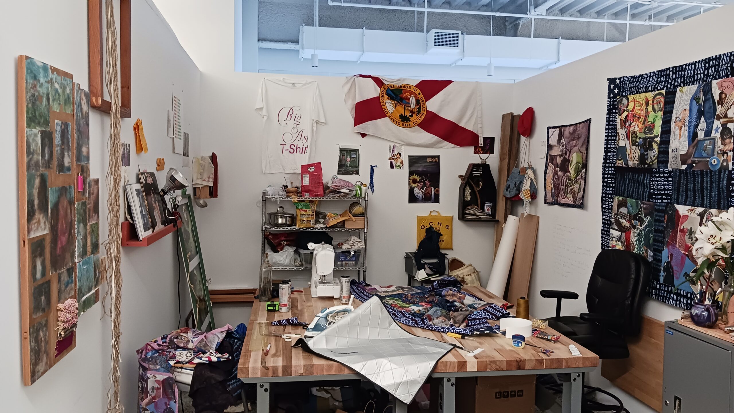 An MFA artist studio at Dock 72. All the walls are covered with various panels, banners, and prints at various scale. A table is in the center of the studio, covered with art supplies.
