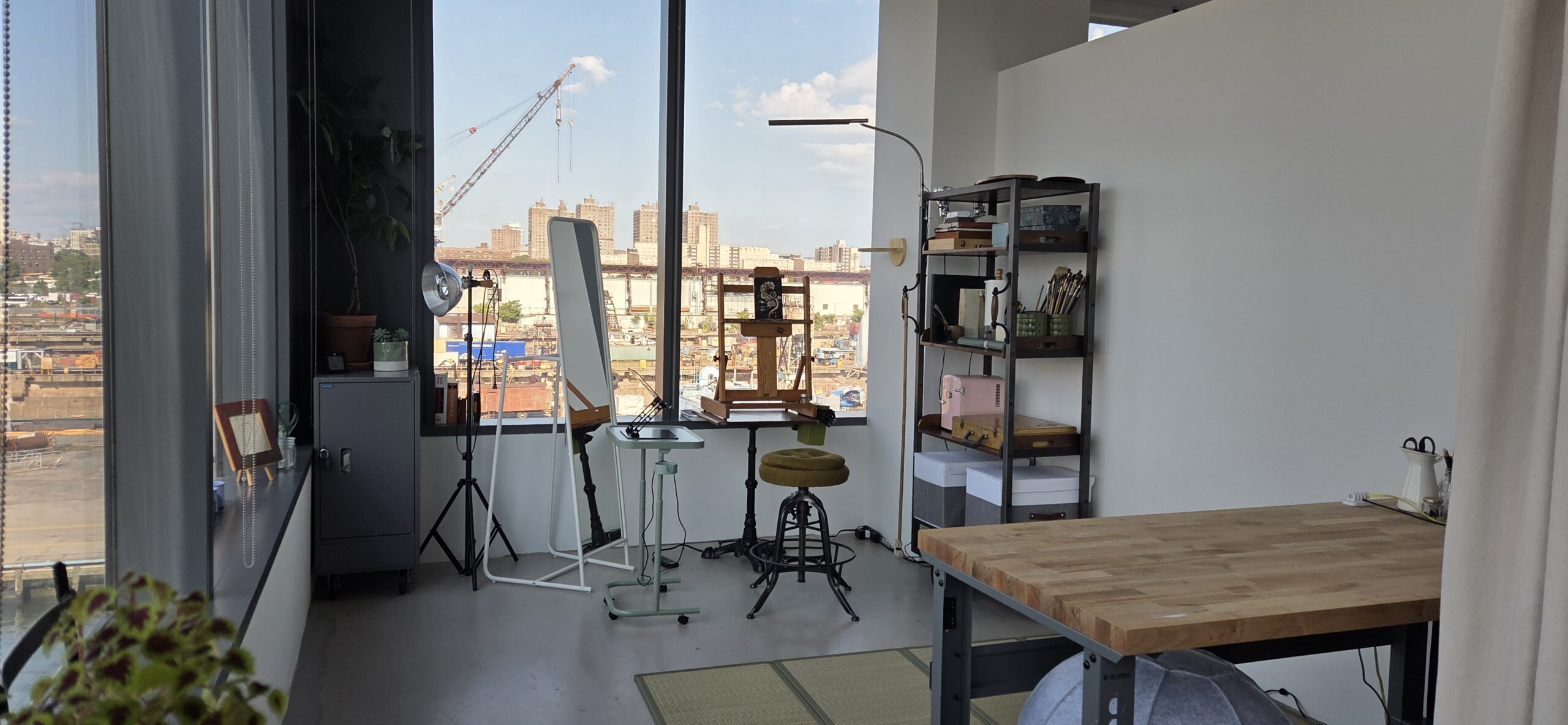 Pratt Fine Arts MFA corner studio in Dock 72. Two windows overlooking the Brooklyn Navy Yard and East River. There is a standing mirror and stool, shelf with supplies, and desk with wood top.