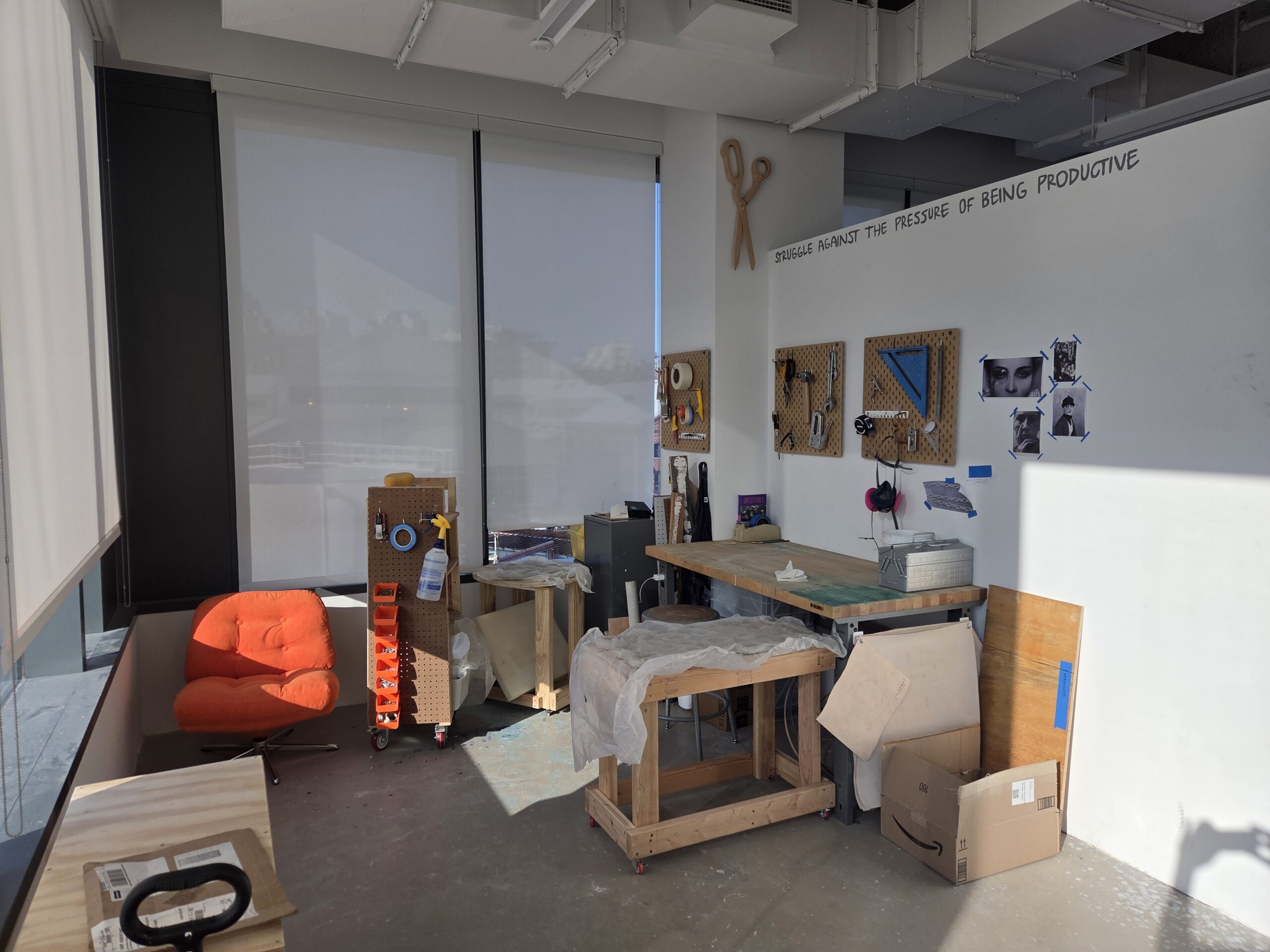 Dock 72 MFA Fine Arts studio. The two corner walls are windows with the shades pulled down. There is a table against each of the opposing walls and a bright orange chair in the back left corner. Small works are hung on the wall above the right-hand table.