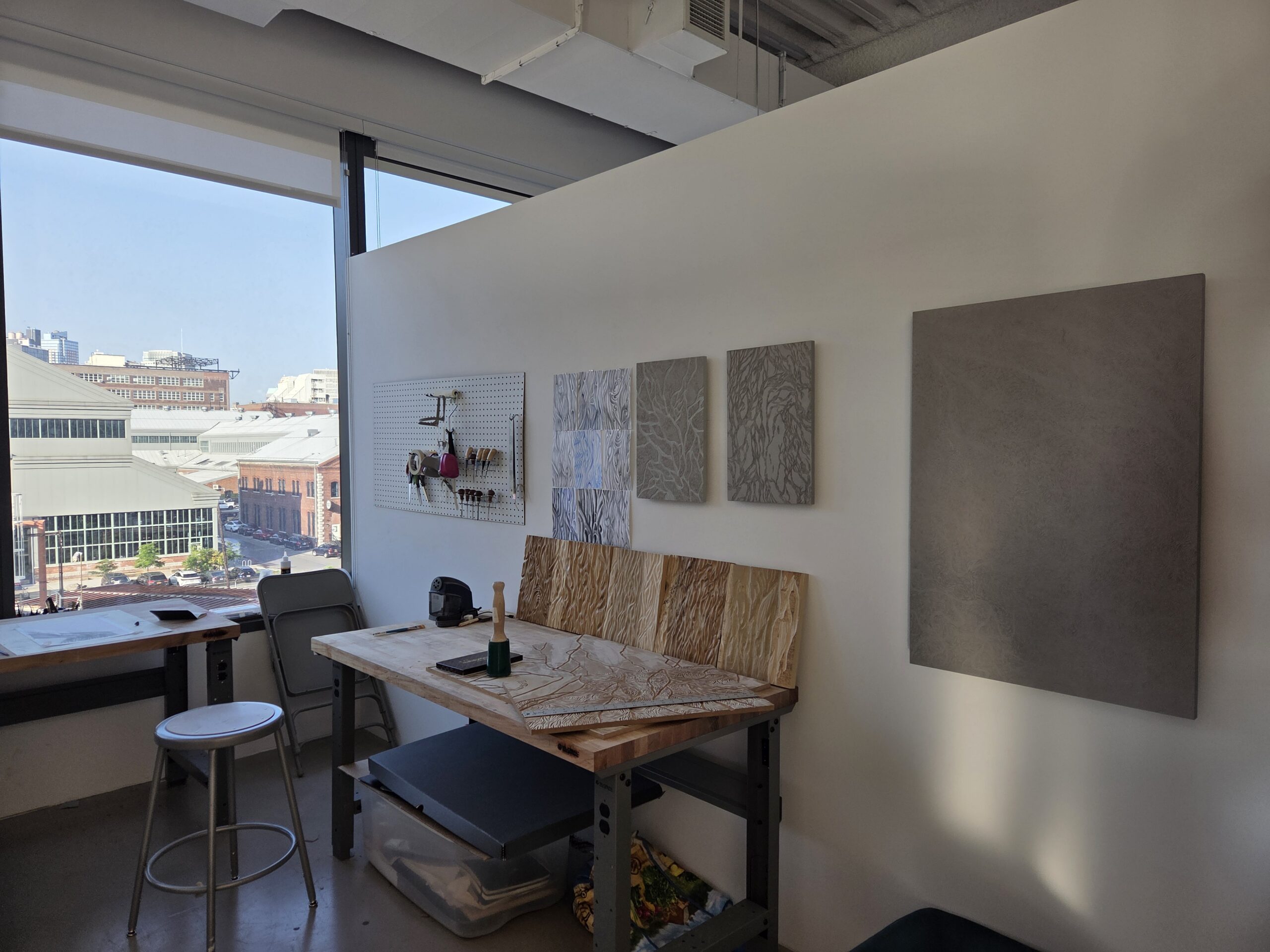 MFA studio at Dock 72 with view of the Brooklyn Navy Yard. Works are hung on the walls, under which there is a table. Leaning against the wall on the table are a series of carved woodblocks.