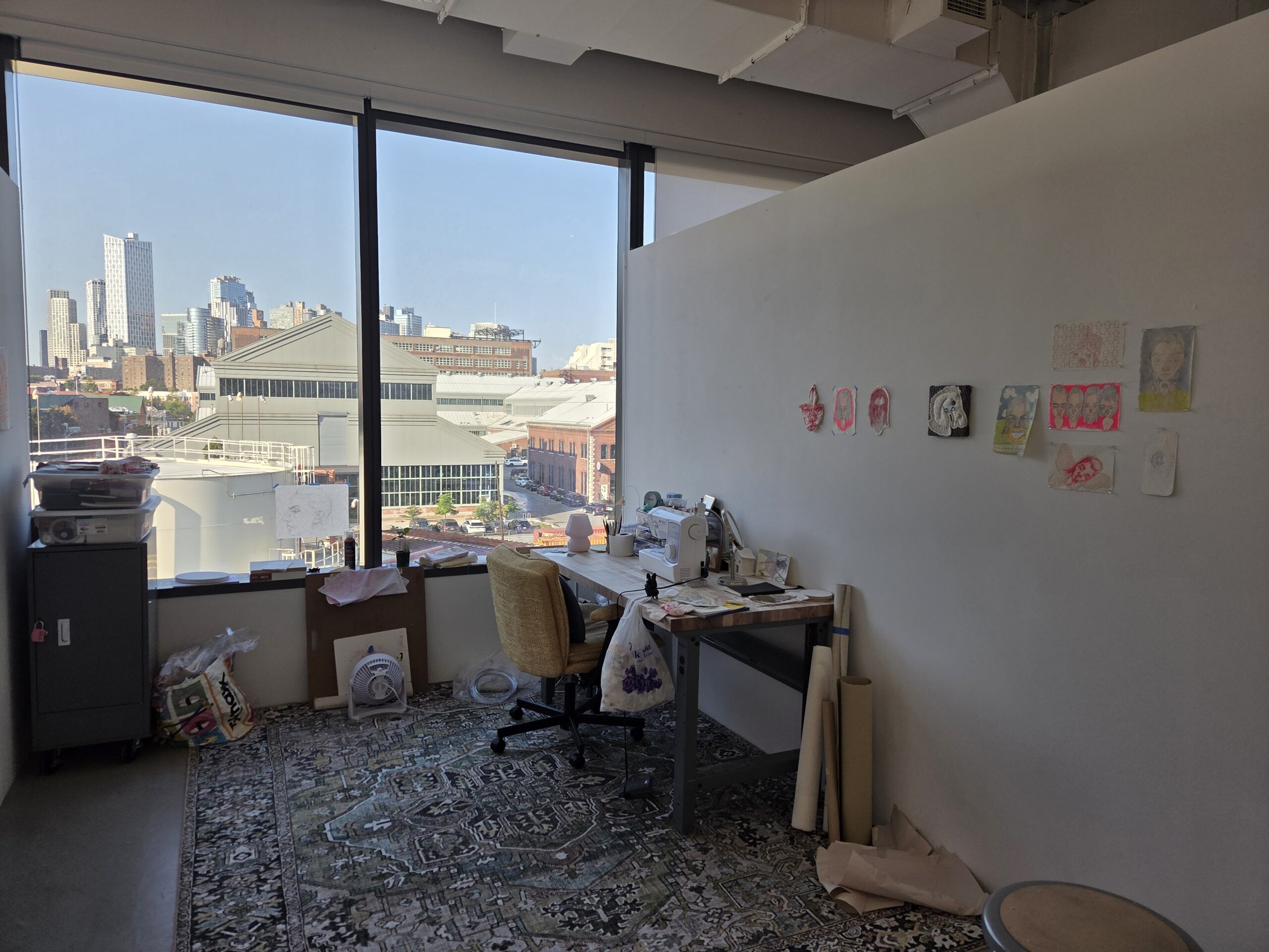 Dock 72 Fine Arts MFA studio with small works installed on the wall. A table is against the right wall with a chair. There is a large rug covering the floor. The window view is of the Brooklyn Navy Yard