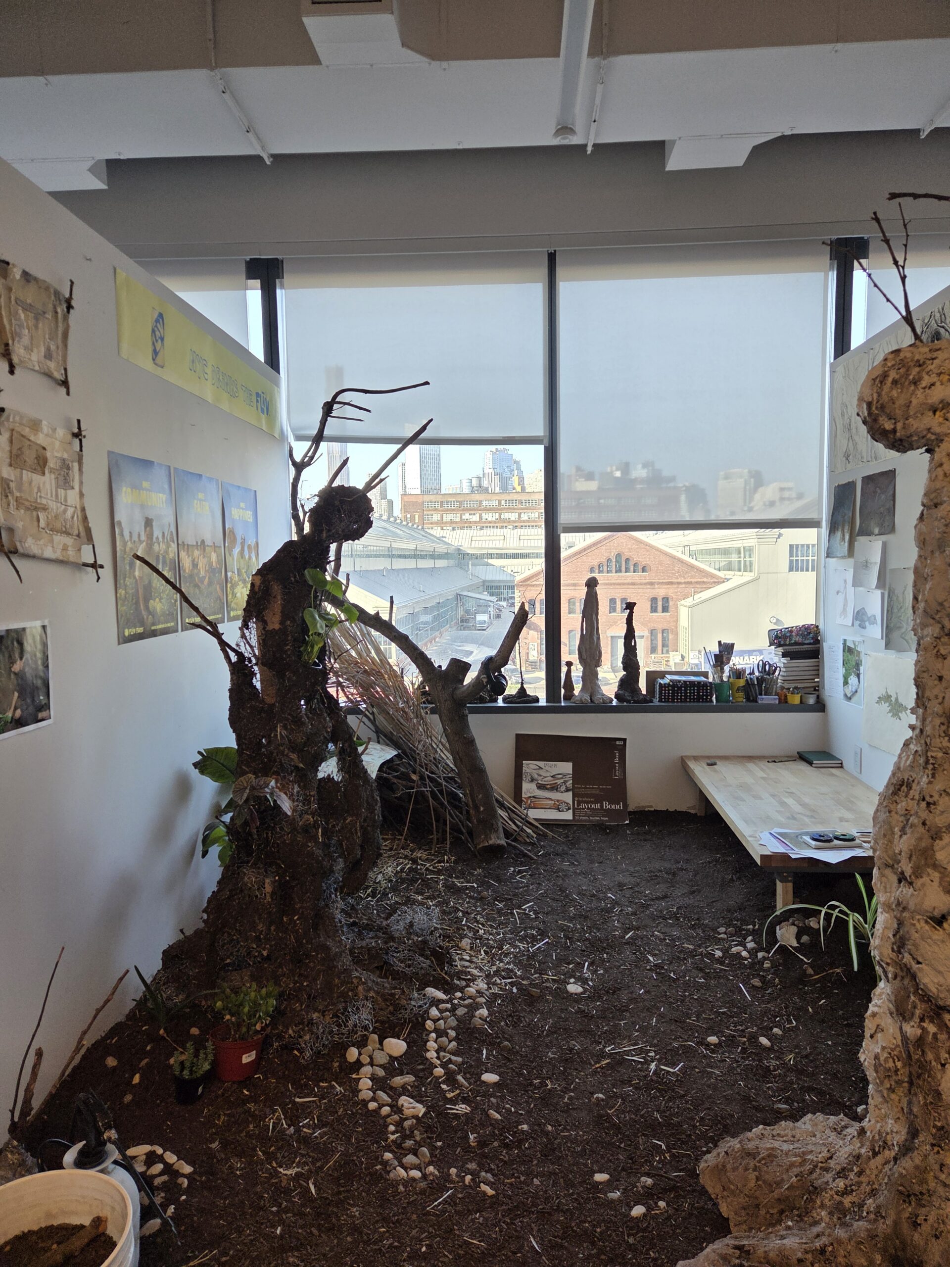 MFA studio at Dock 72. Soil covers the entire floor of the studio, and a tree-like piece of wood stands toward the left wall. Various posters and other artworks are hanging on the wall. The window overlooks the Brooklyn Navy Yard
