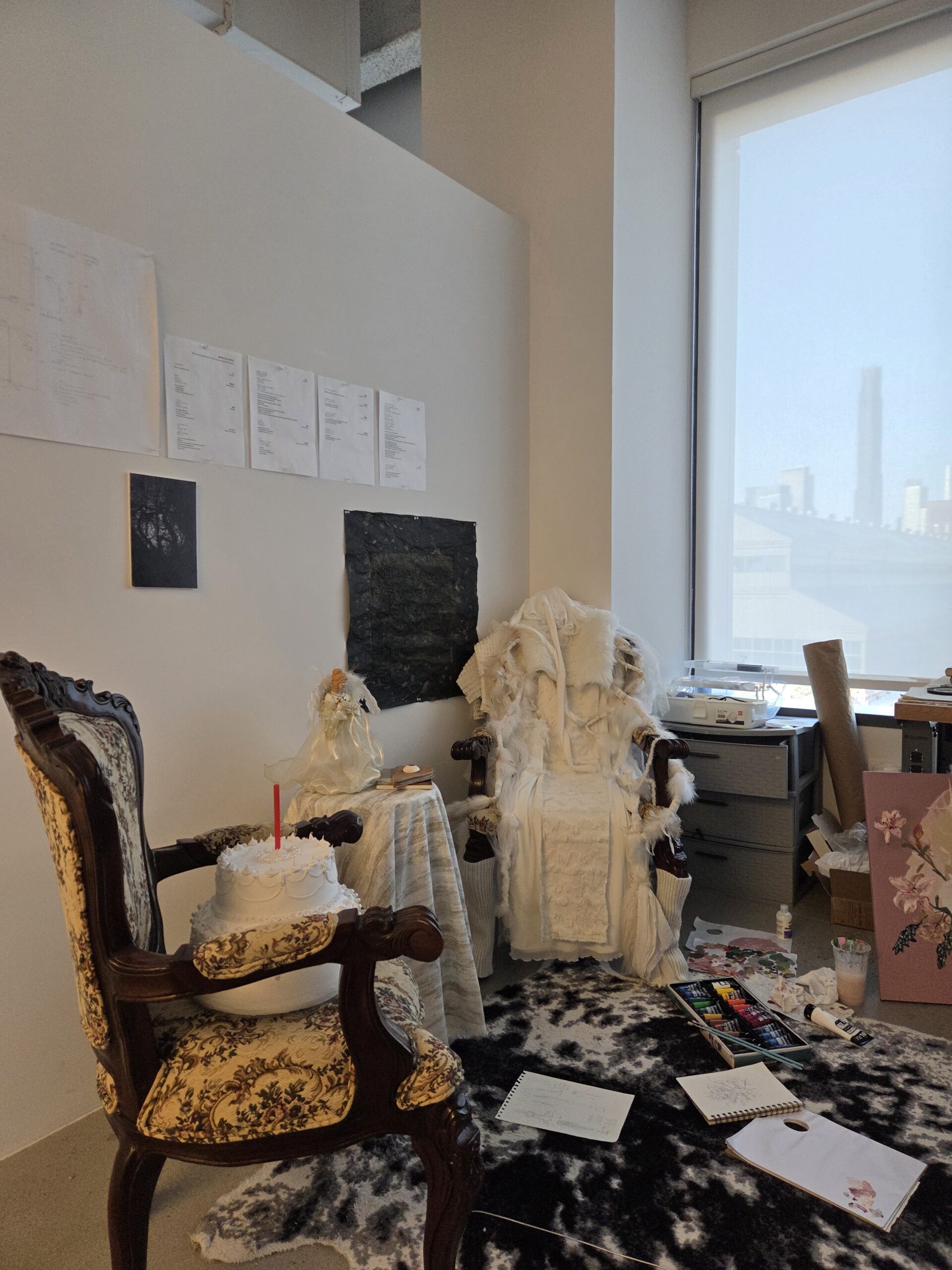 Fine Arts MFA studio at Dock 72. Two chairs, one with a cake on the seat, the other with a fiber piece draped over it. Small works are pinned to the wall. The floor is covered by a rug and various art supplies