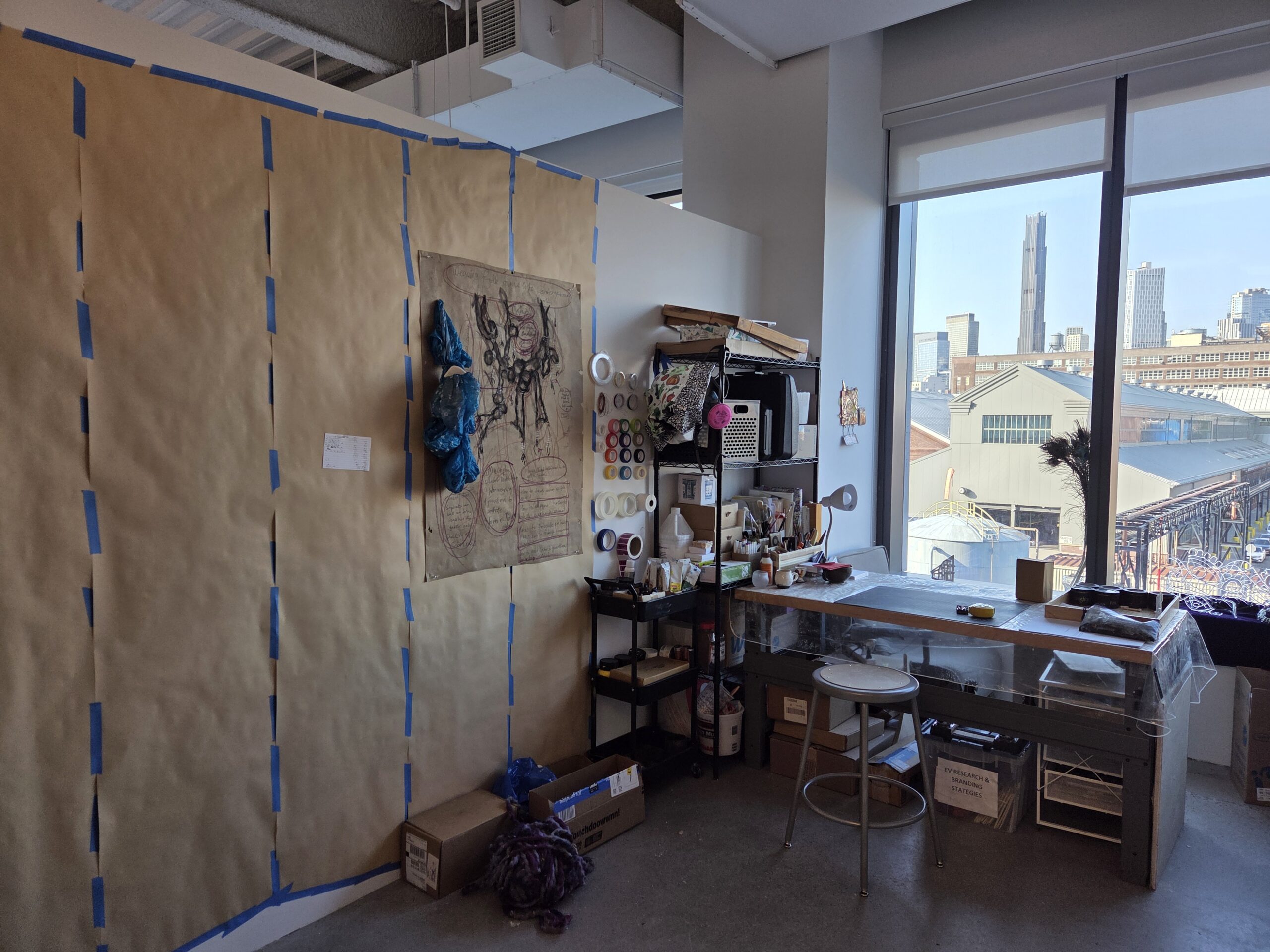 MFA studio at Dock 72 with view of the Brooklyn Navy Yard. Large pieces of brown paper are taped to the wall. There is also a desk and various art supplies.