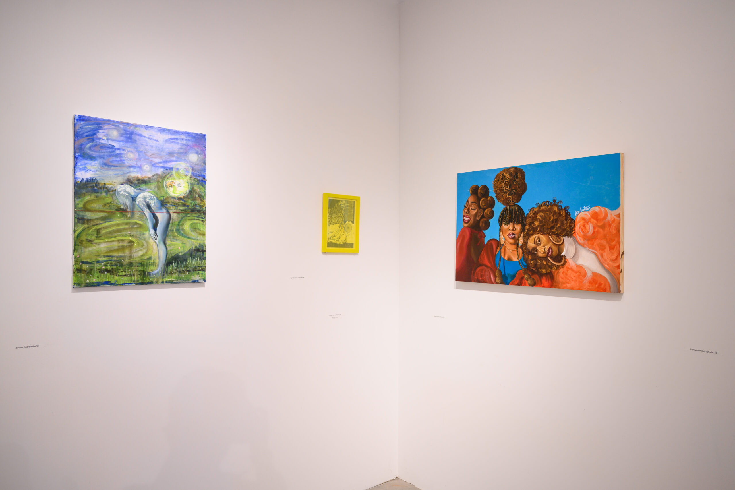 A gallery corner with works installed on the walls. Left to right: a small panel with blue and green, a small yellow work, and a painting of a group of people in bright blue and orange and brown.