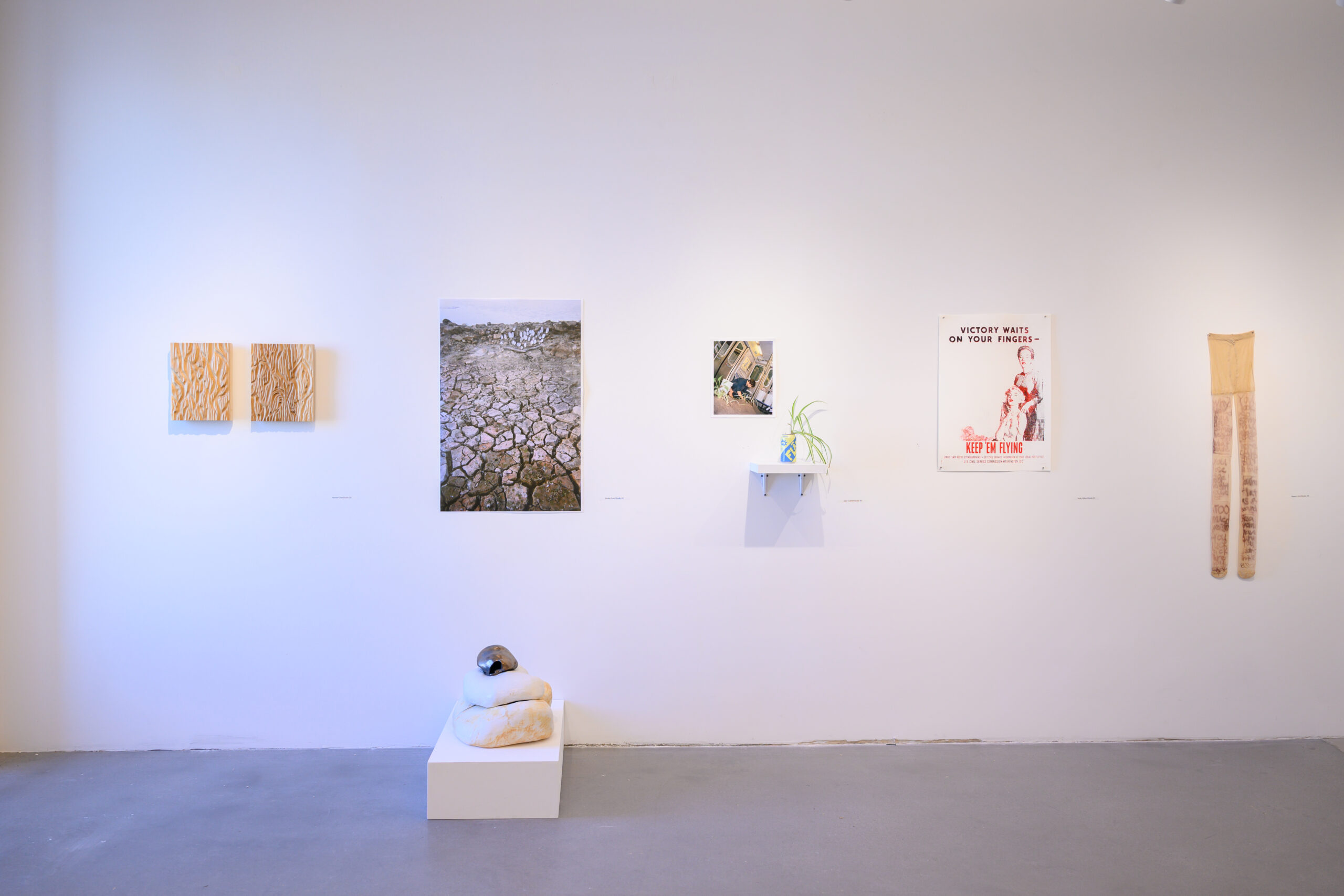 White wall with objects installed. One is a pair of nylon tights. On the floor in front of the wall is a small, low pedestal with a sculpture on it.