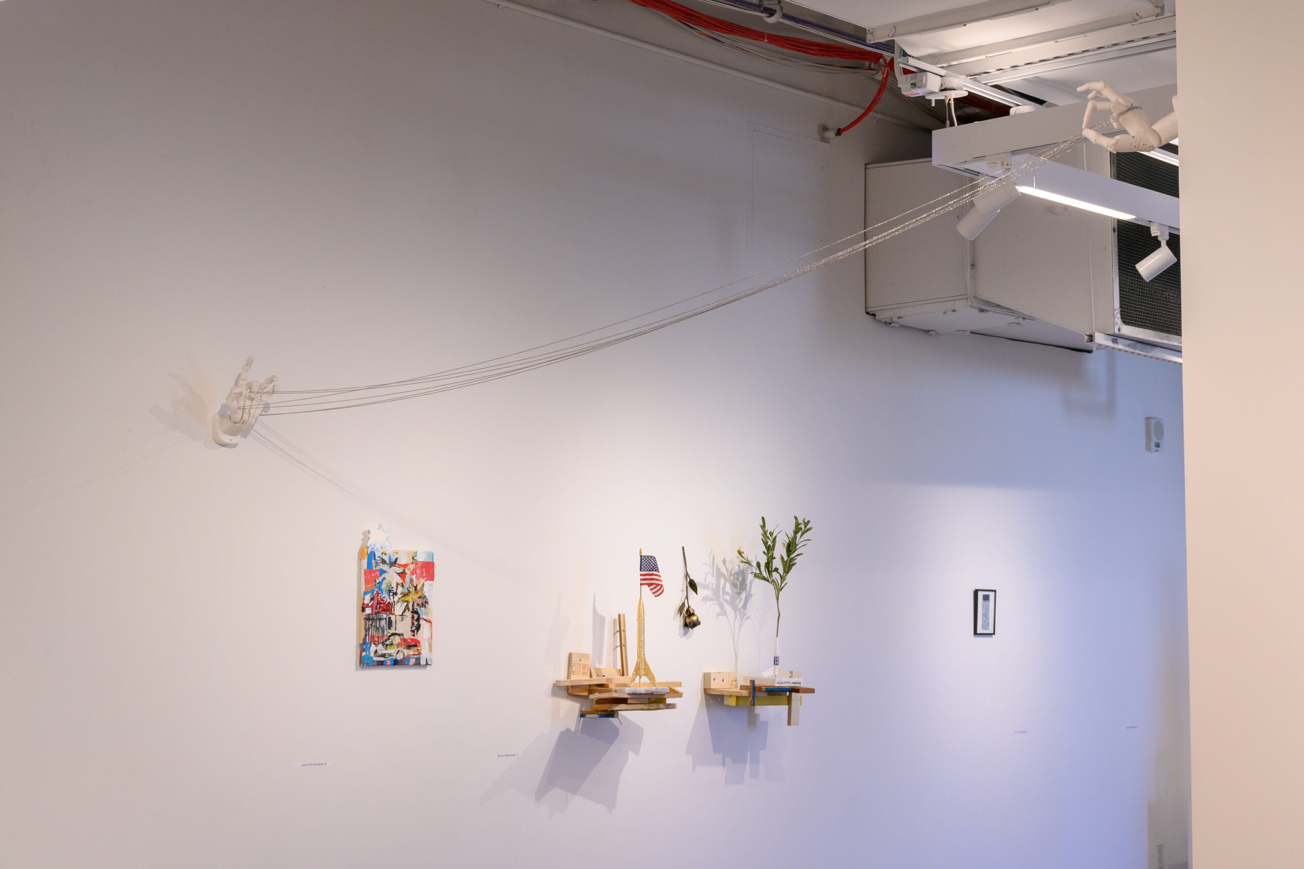 Gallery corridor installation. A small work and two small shelves with sculptures are installed on the wall. From wall to wall are two hands installed high up, connected by chains.