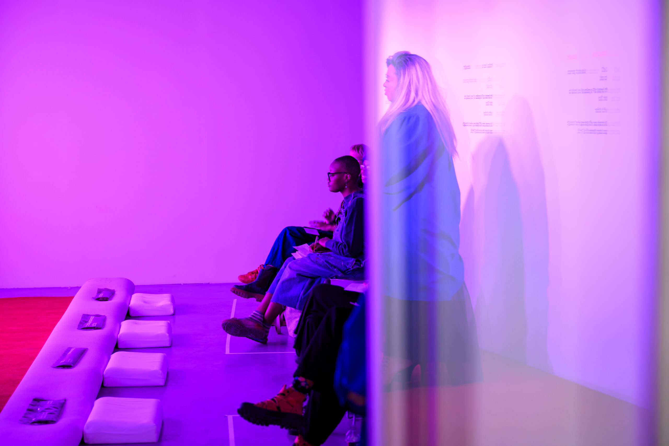 A red carpet laid on the floor with a long pillow and kneeling cushions. A row of people are seated facing toward where the carpet and cushions are situated, with one person standing with a long blue robe on. Behind them is text on the wall. The lighting in the room is purple.