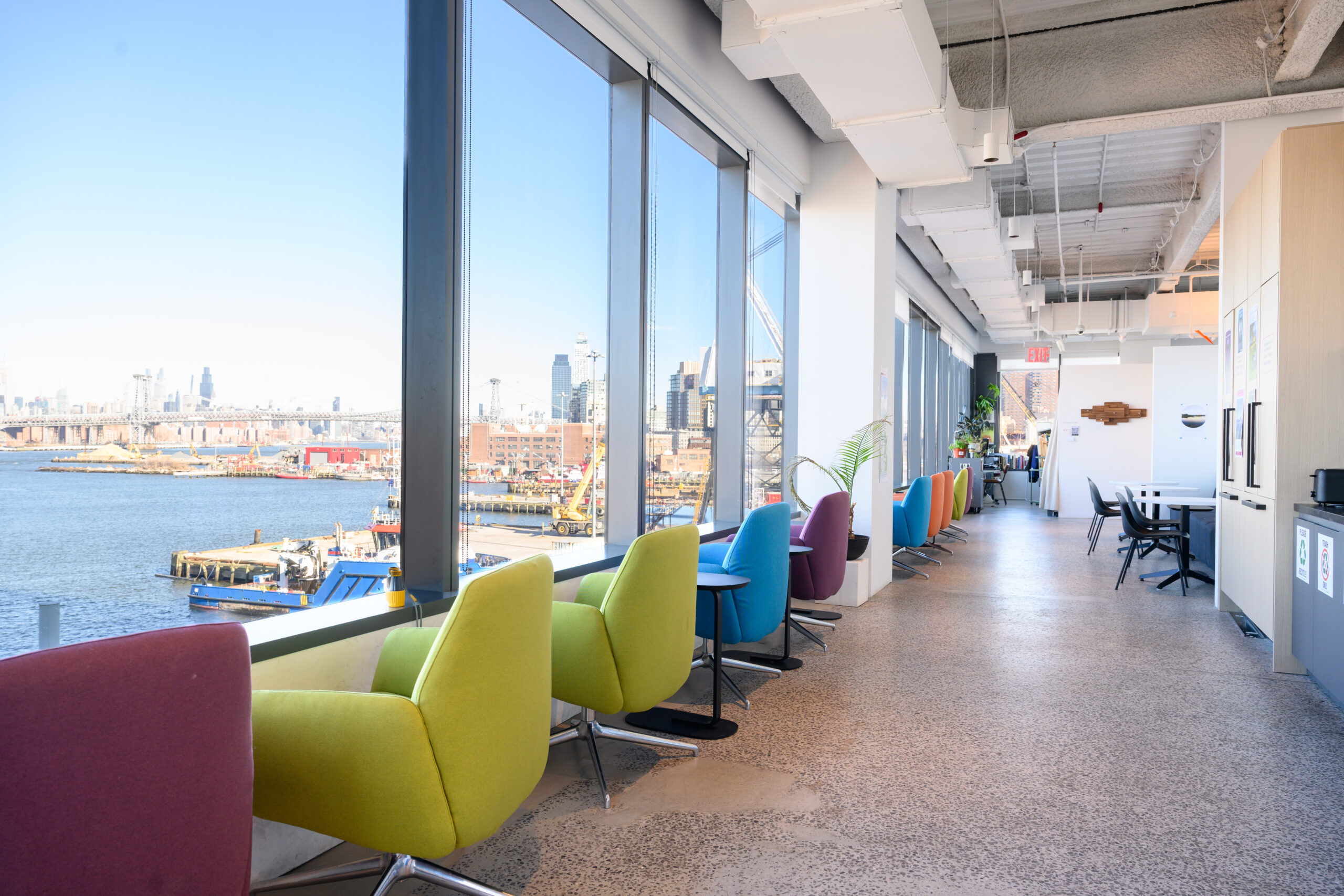 Lounge area in the Dock 72 space. A row of colorful cushioned chairs in a row facing a large window overlooking the East River