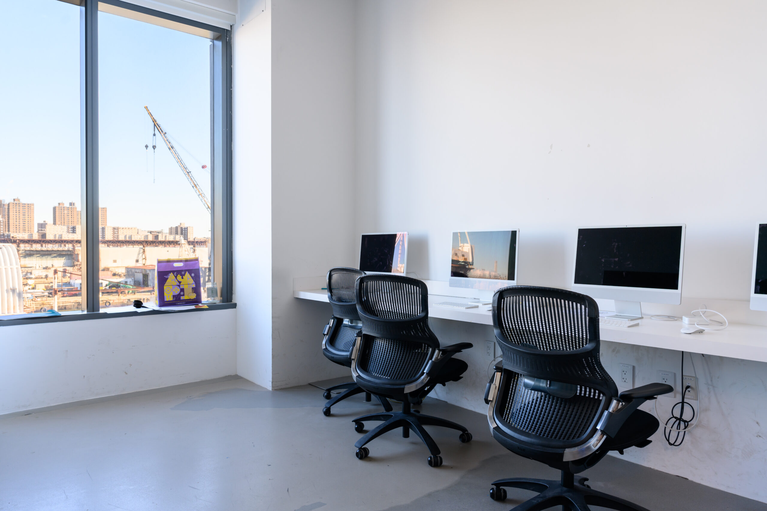 Dock 72 computer lab. A row of Mac monitors with rolling chairs. The window shows the Brooklyn Navy Yard.