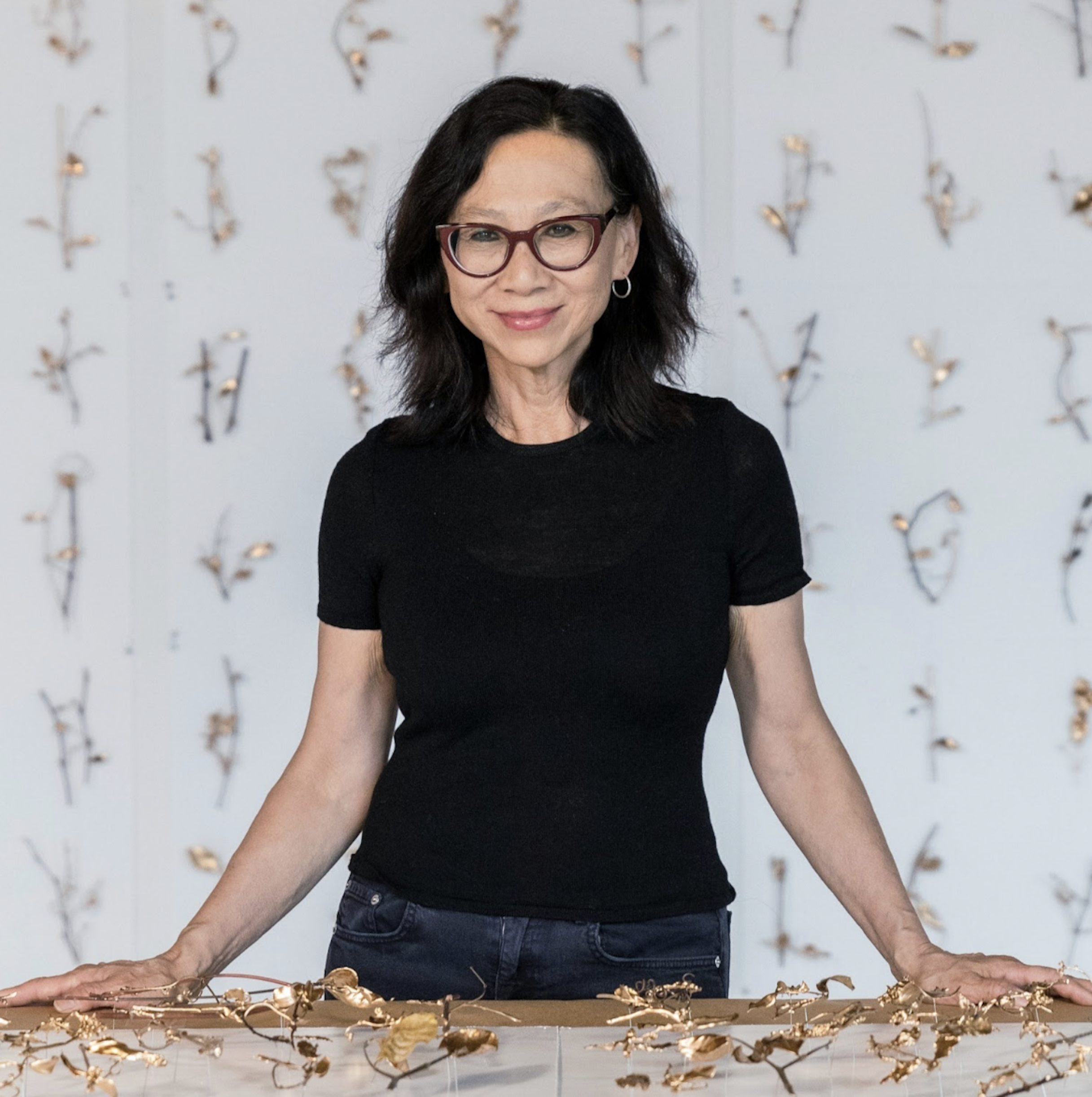 Portrait of Yu-Wen Wu in center of the frame wearing a black t-shirt and dark-rimmed glasses. She rests her hands on a table in front of her. Behind here are small works installed on the wall in a grid.