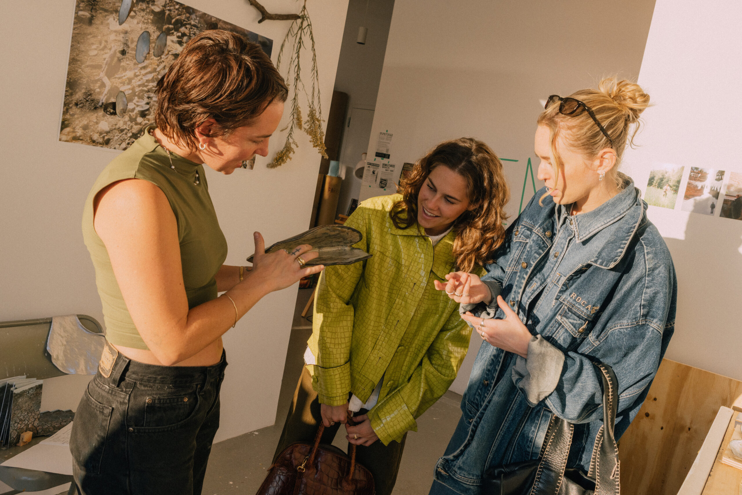 Three women are engaged in conversation in a bright, art-filled space. One woman, wearing a green top, is showing an object to the other two, who are attentively listening. One of the listeners is dressed in a bright yellow jacket, while the other wears a denim jacket. Artwork and materials are visible on the walls and workspace, reflecting a creative environment.