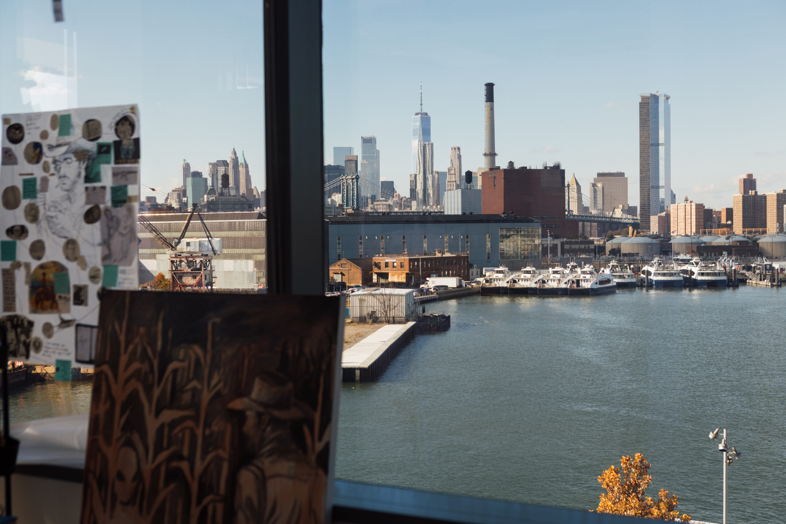 A view from a window showcases a waterfront scene with boats docked along a pier. In the background, a skyline features tall buildings, including skyscrapers. Two artworks are positioned in the foreground: one is a collage, and the other is a painting depicting a figure in a hat in a field. The scene is well-lit with clear skies.