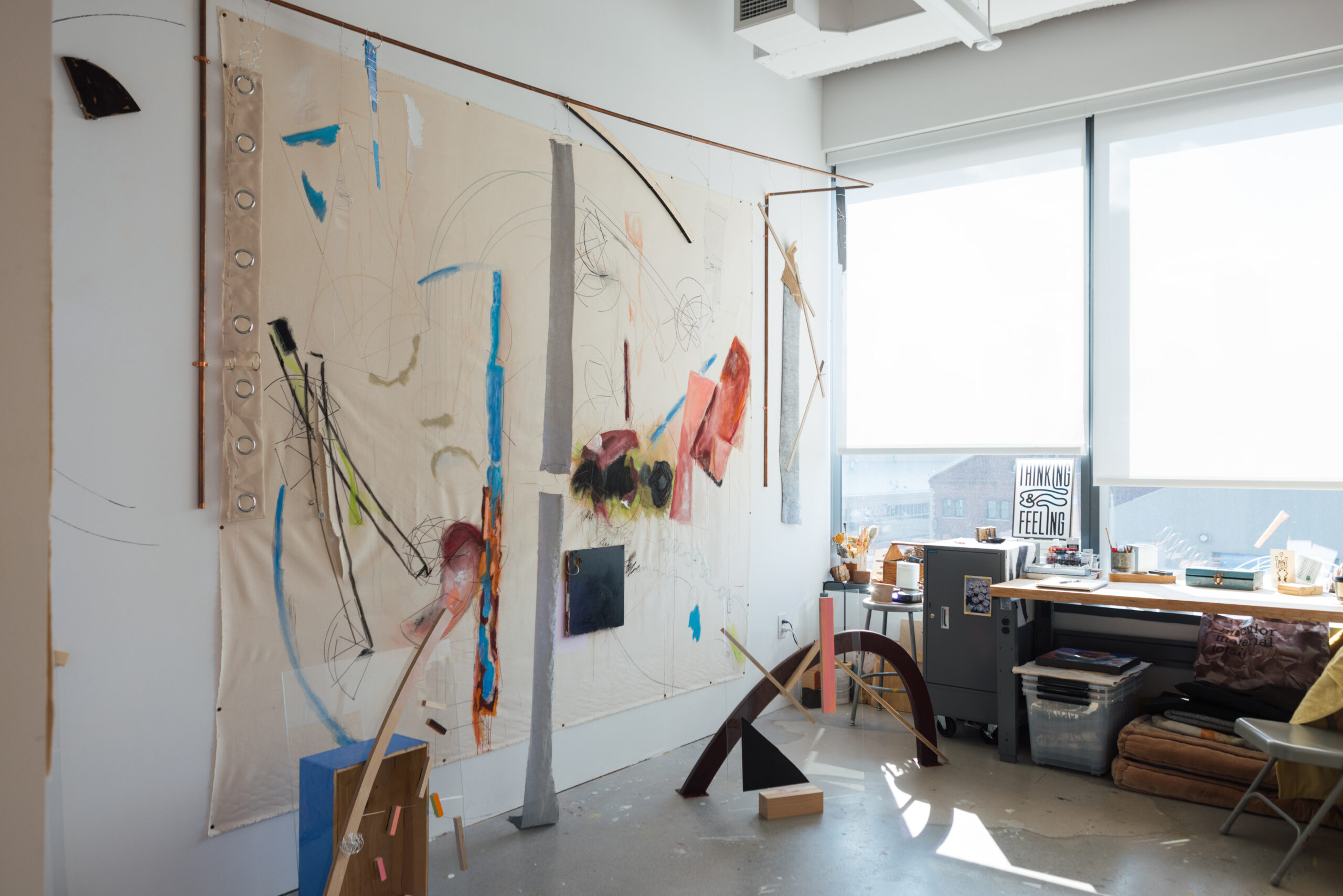 A bright artist's studio features a large abstract artwork mounted on the wall, filled with various colors and shapes. In the foreground, several geometric wooden and acrylic sculptures are positioned on the floor. A worktable against the opposite wall holds art supplies, a framed poster with the text "THINKING & FEELING," and other materials, while sunlight streams in through large windows, illuminating the space.