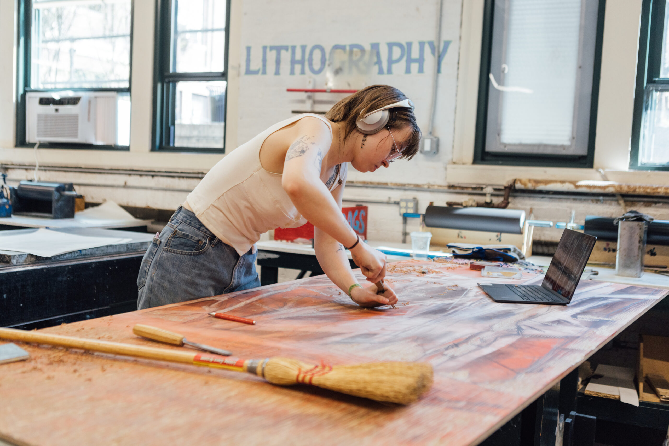 A person with short brown hair is working intently at a large wooden table covered in art materials. They are wearing headphones and are using a carving tool on a piece of lithography stone. There are art supplies and a laptop on the table, with some shavings visible around the workspace. The background features large windows and the word "LITHOGRAPHY" painted on the wall.