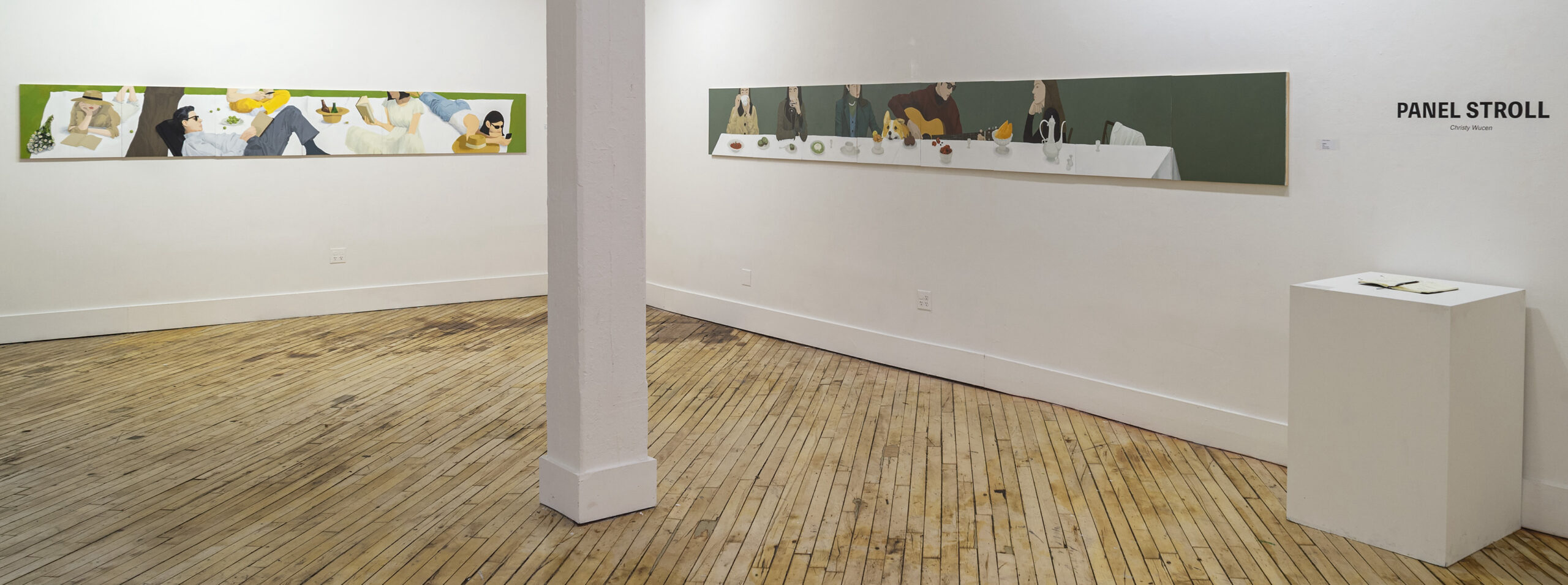 An art gallery features two large, colorful murals on the walls, depicting people at gatherings and a table set with food. A white pedestal displays a notebook and is labeled "PANEL STROLL" with the artist's name, Christy Wucen. The floor is made of wooden planks, and there is a structural column in the center of the room. The walls are painted white, highlighting the artwork.