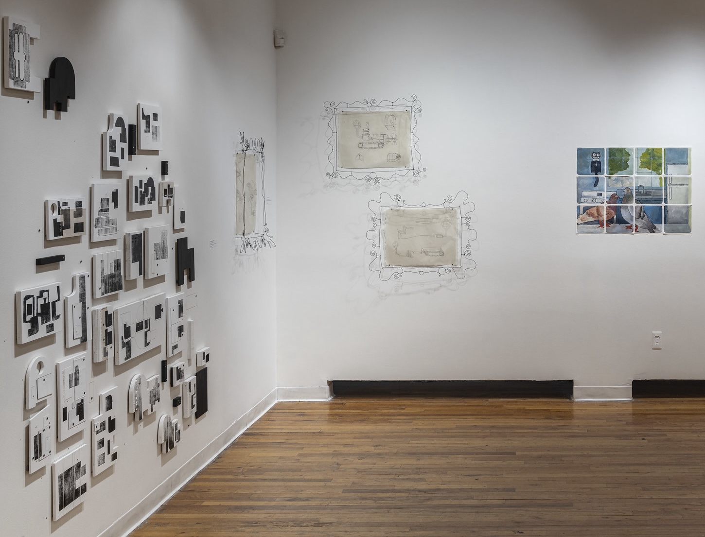 The image shows an exhibition space featuring various artworks. On the left wall, there are multiple framed pieces in black and white, arranged in a collage pattern. The right wall displays two outlined frames containing sketches and a grid of watercolor images depicting urban scenes with birds and figures. The floor is wooden, and the overall ambiance is minimalist and artistic.