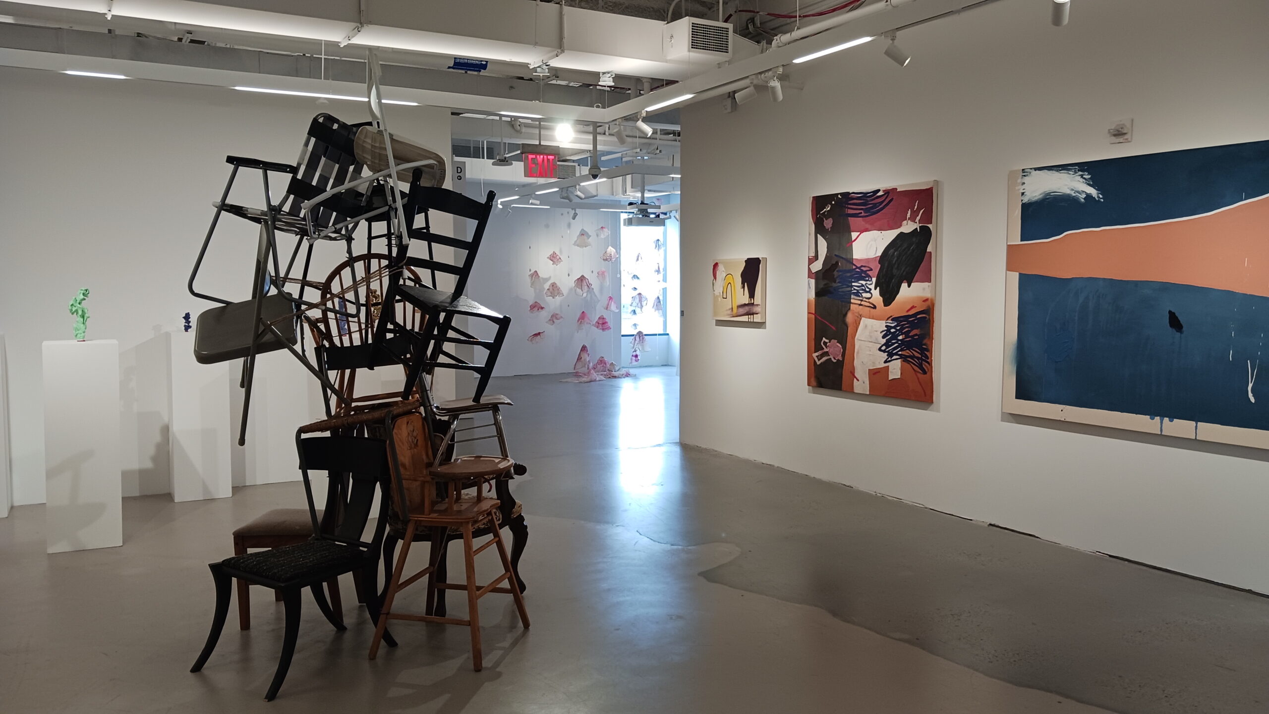 An art gallery space featuring a sculpture made of stacked chairs in the foreground. To the right, two colorful paintings hang on the wall. In the background, delicate fabric sculptures are suspended from the ceiling, displaying soft colors. There are also white pedestals displaying small, possibly sculptural artworks. Natural light comes in from a large window at the end of the hallway.