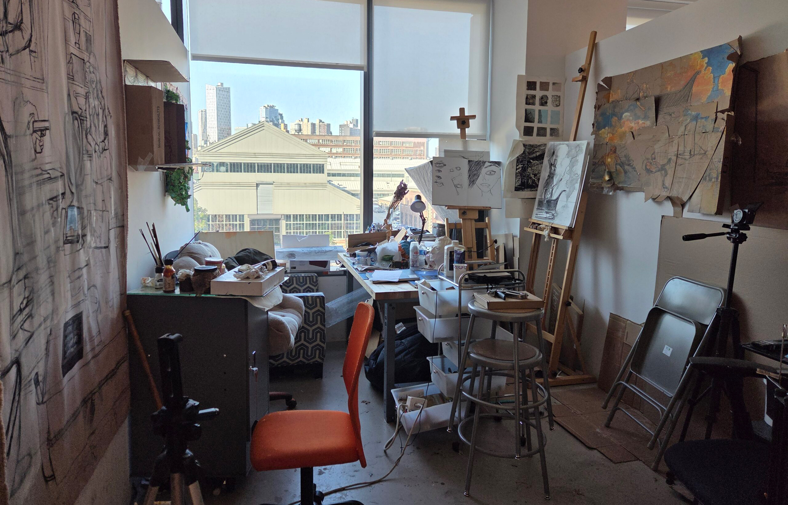 A cluttered artist's studio with an orange chair in the foreground. The workspace includes unfinished artwork on easels, sketches pinned to the wall, various art supplies, and a large window revealing a cityscape outside. Additional furniture such as a gray filing cabinet, stools, and an outdoor view of buildings are also visible. Natural light streams into the room.
