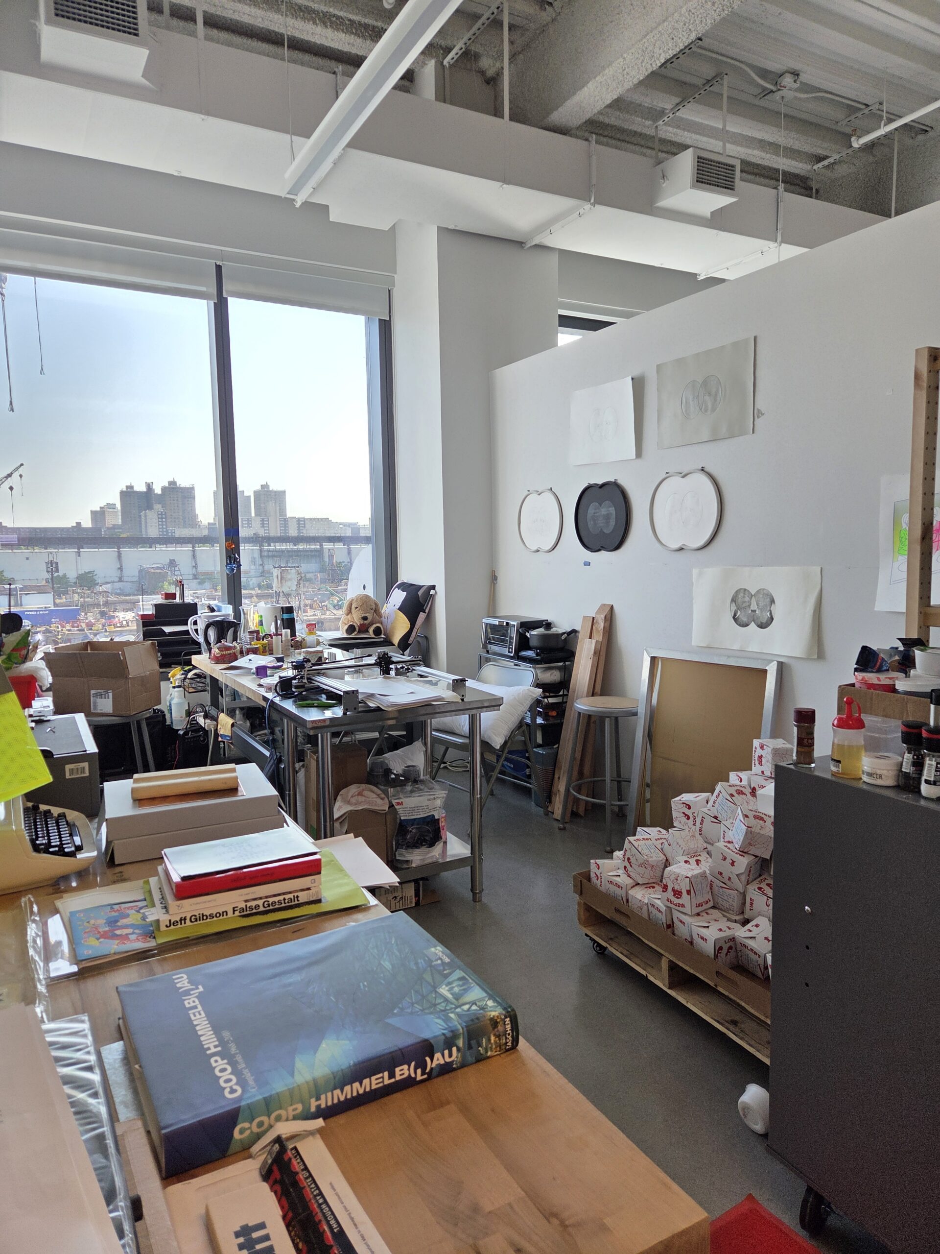 A bright, open studio space with large windows showing a cityscape. The room has various art supplies, books, and paperwork scattered on tables, along with stacks of takeout containers on a cart. On the wall, there are sketch-like artworks displayed. The flooring appears to be concrete, and there are items organized around the workspace, suggesting a creative environment.