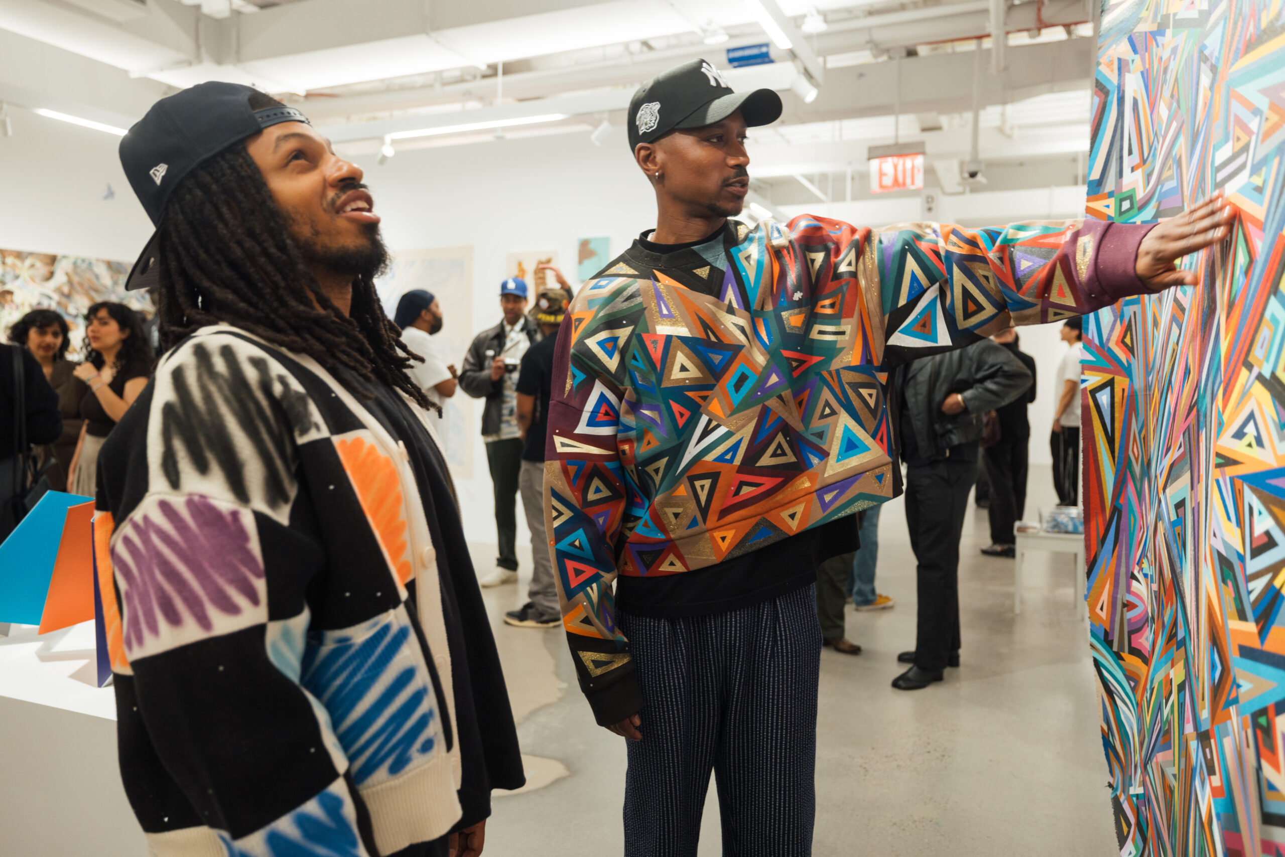 Two individuals are engaged at an art gallery. One has long dreadlocks and wears a patterned black, white, and colorful sweater, with a playful smile as they look towards the other. The second person wears a brightly colored geometric-patterned cropped jacket and is gesturing towards an abstract artwork on the wall. In the background, several other attendees are also observing the art.