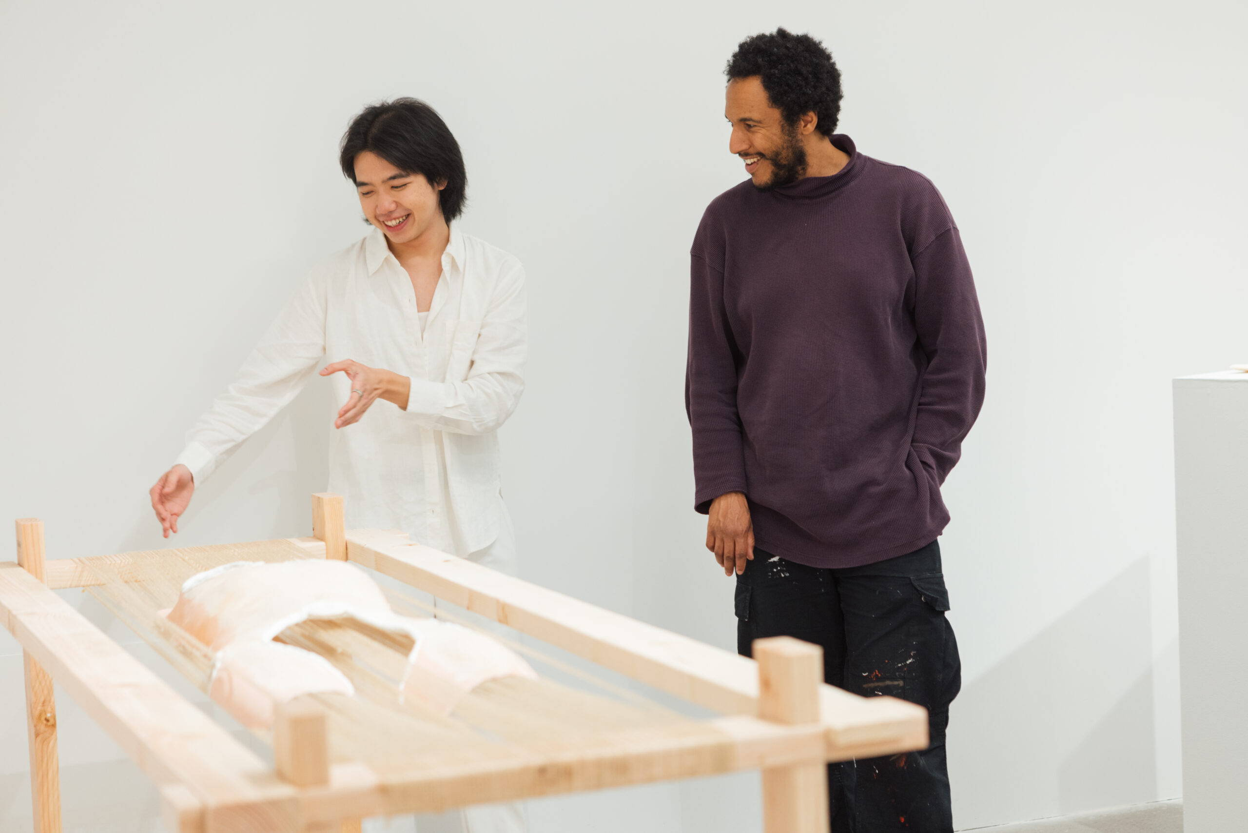 Two people are engaged in conversation in an art gallery. One person, wearing a white shirt, gestures toward an artwork displayed on a wooden table, while the other, dressed in a dark purple sweater, smiles. The background is a plain white wall, and there is a small pedestal with another piece of art visible on the right.