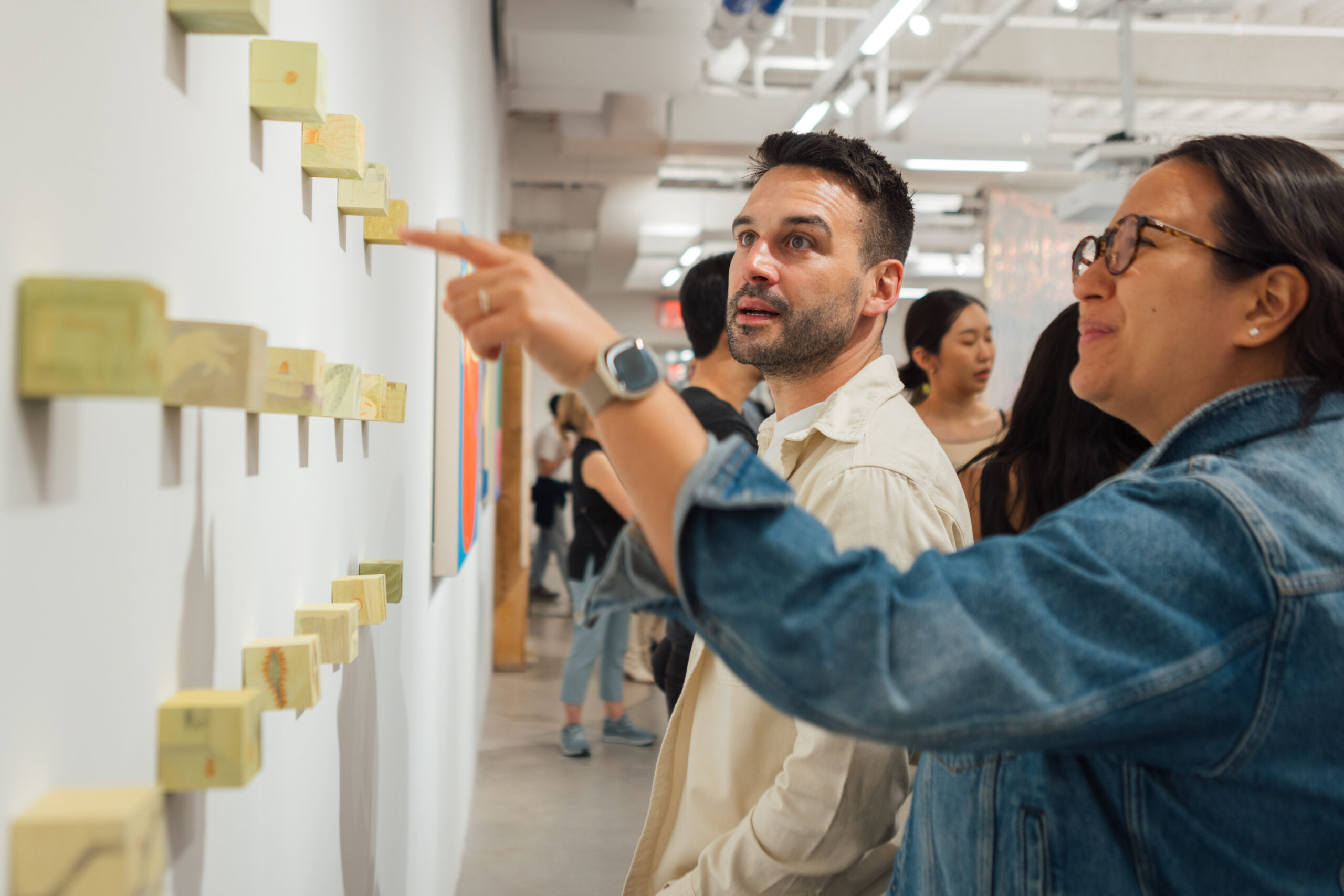 A man and a woman are interacting in an art gallery, looking at a series of small wooden artworks mounted on a wall. The man, wearing a light shirt, appears engaged as he looks at the pieces, while the woman, in a denim jacket, points to one of the artworks with a smile. In the background, other visitors stroll through the gallery.