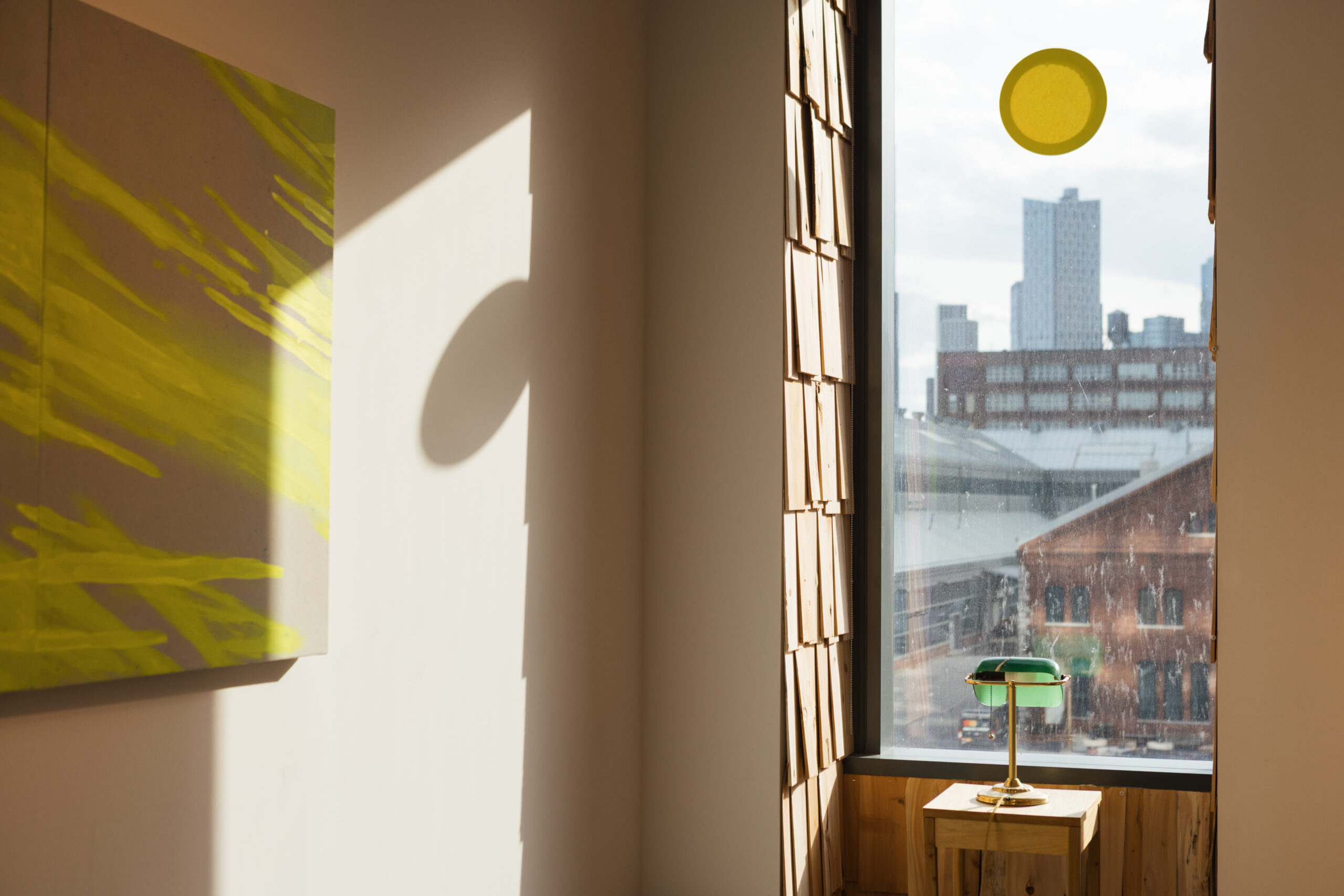 A bright corner of a room features a large window with skyline views, partially blocked by a circular yellow decoration. Sunlight streams in, casting shadows against the wall, which displays an abstract painting with vibrant yellow streaks. A green and gold lamp sits atop a wooden table beneath the window, adding warmth to the space. Wooden panels line the wall, enhancing the natural aesthetic.