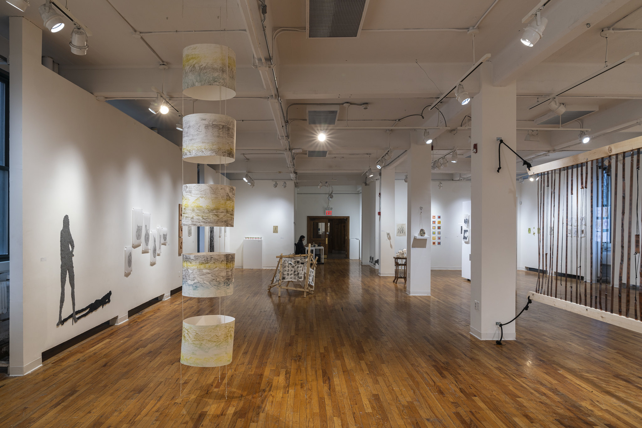 An art gallery interior with wooden flooring and high ceilings. Suspended from the ceiling are three cylindrical fabric pieces featuring colorful designs. On the walls, various artworks, including a silhouette of a figure and framed pieces, are displayed. In the background, a person sits at a table. The space is well-lit with track lighting, creating a bright atmosphere for the exhibition.