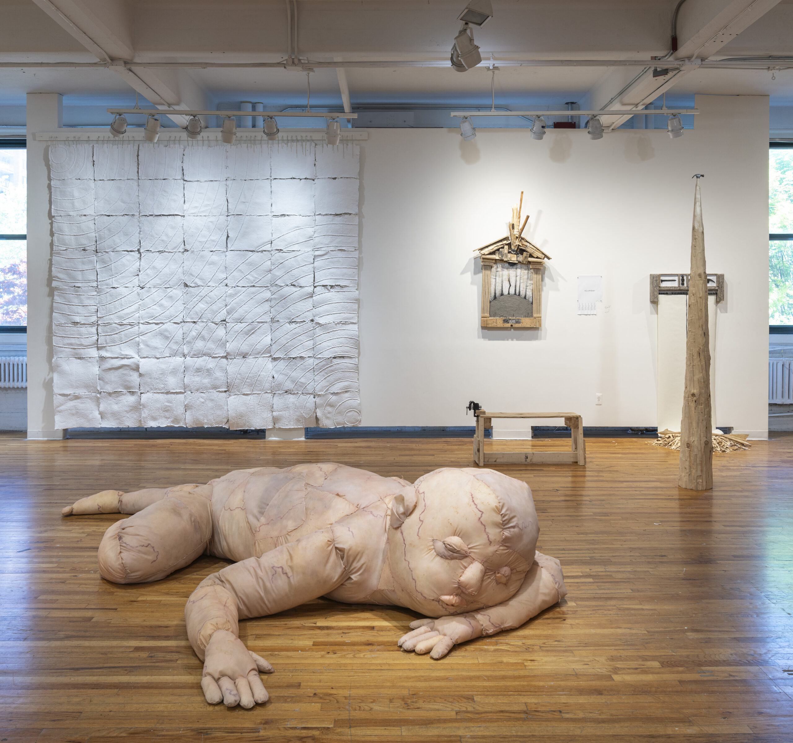 A spacious art gallery featuring a large, soft sculpture of a humanoid figure lying on the wooden floor. The figure has a pale, textured surface resembling skin. In the background, there is an abstract white wall piece made of squares with varying textures and curves. Other artworks include a wooden structure with sticks, a tall wooden figure, and a small table. Natural light filters in through a window, illuminating the space.