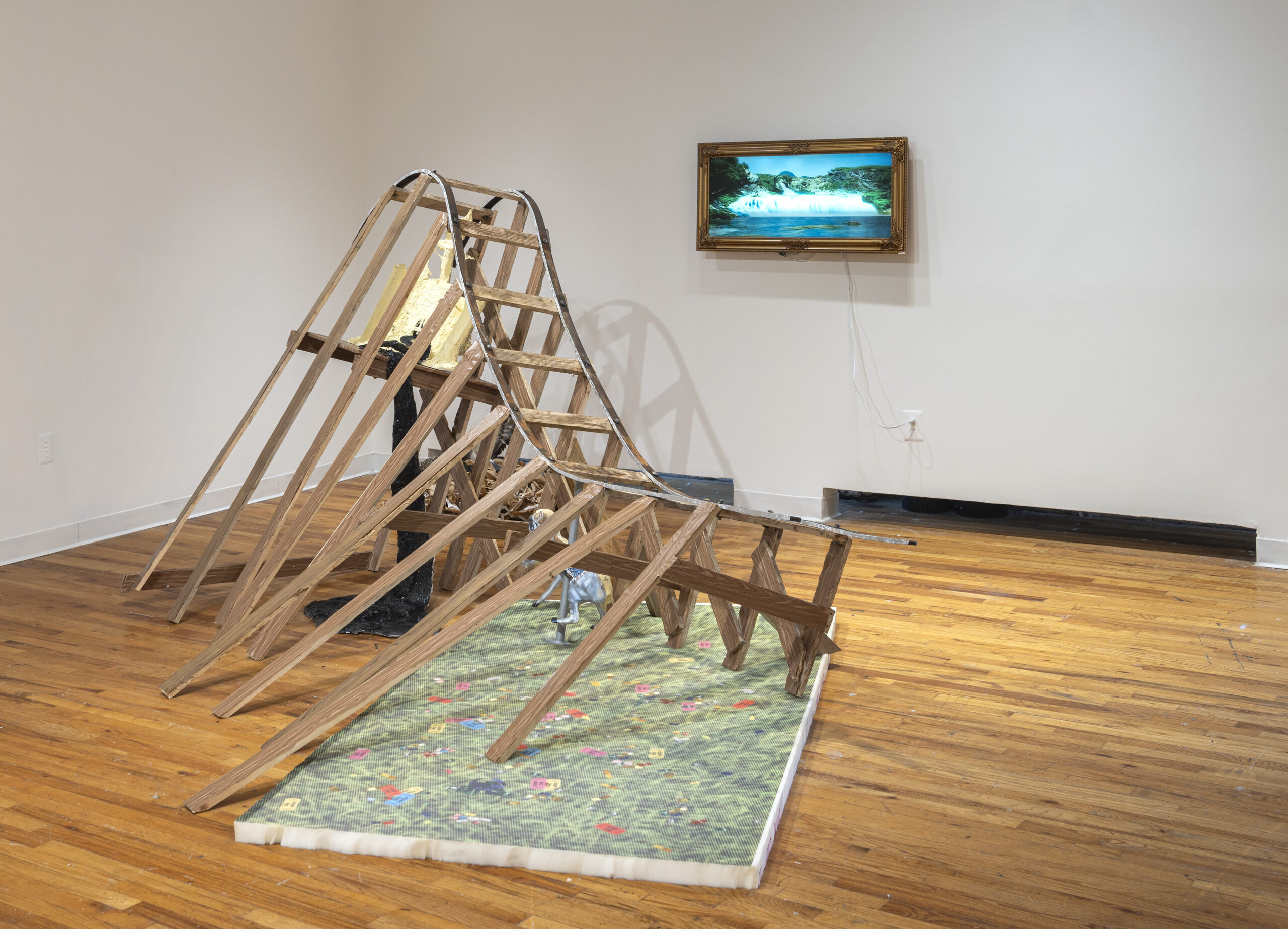 An art installation features a wooden roller coaster-like structure partially set on a colorful grassy mat with floral patterns. A small sculpture appears near the base of the structure. On the wall behind it is an ornate, framed television displaying a serene landscape scene. The floor is made of wooden planks, and the space has minimal decoration.