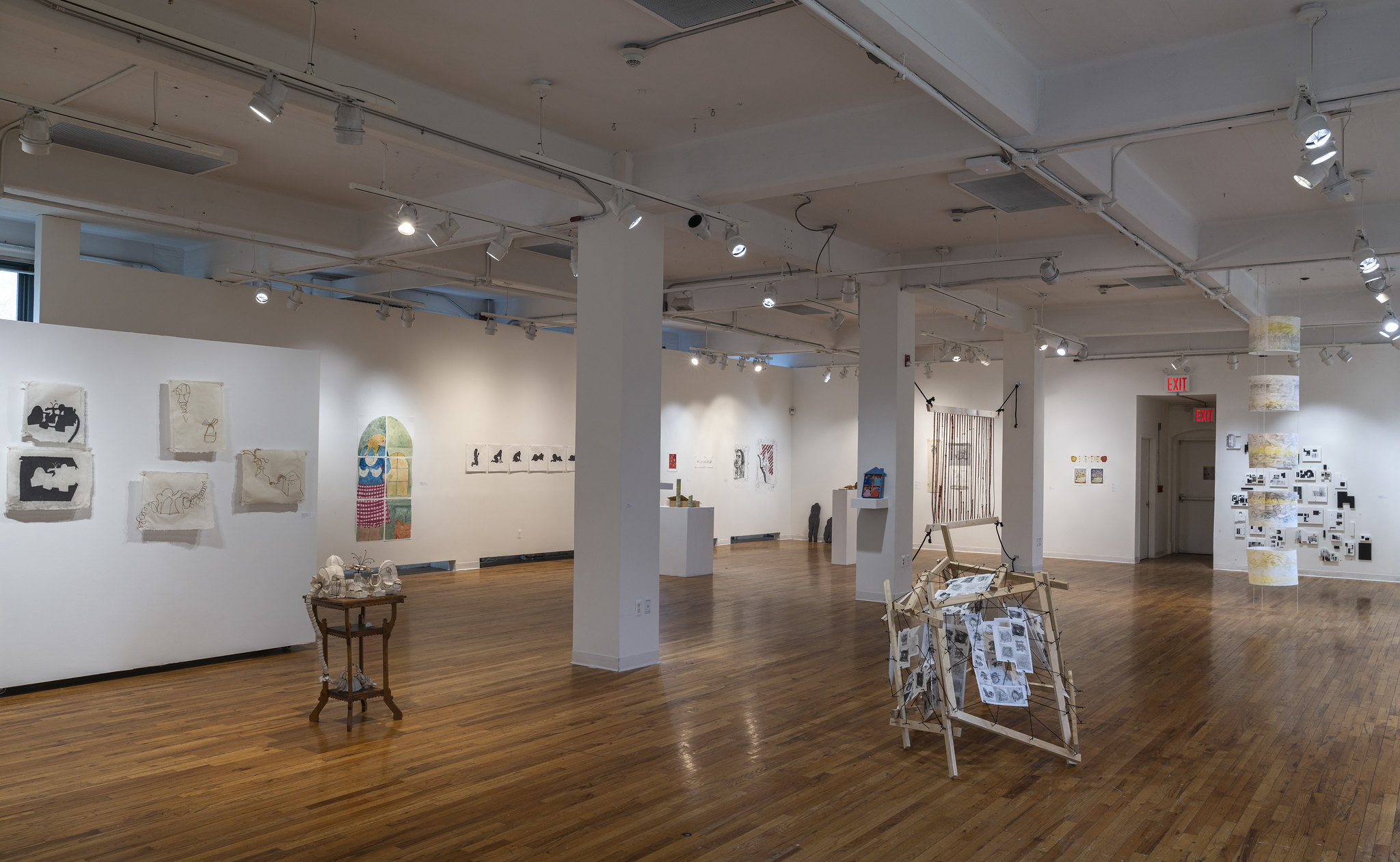 An art gallery interior featuring wooden floors and white walls. Various artworks are displayed on the walls, including framed images, paper works, and a colorful archway. In the center, there is a wooden cart with unique objects, and an installation made from wooden sticks with printed pages hanging. Artworks cover different styles and mediums throughout the space, illuminated by adjustable ceiling lights.