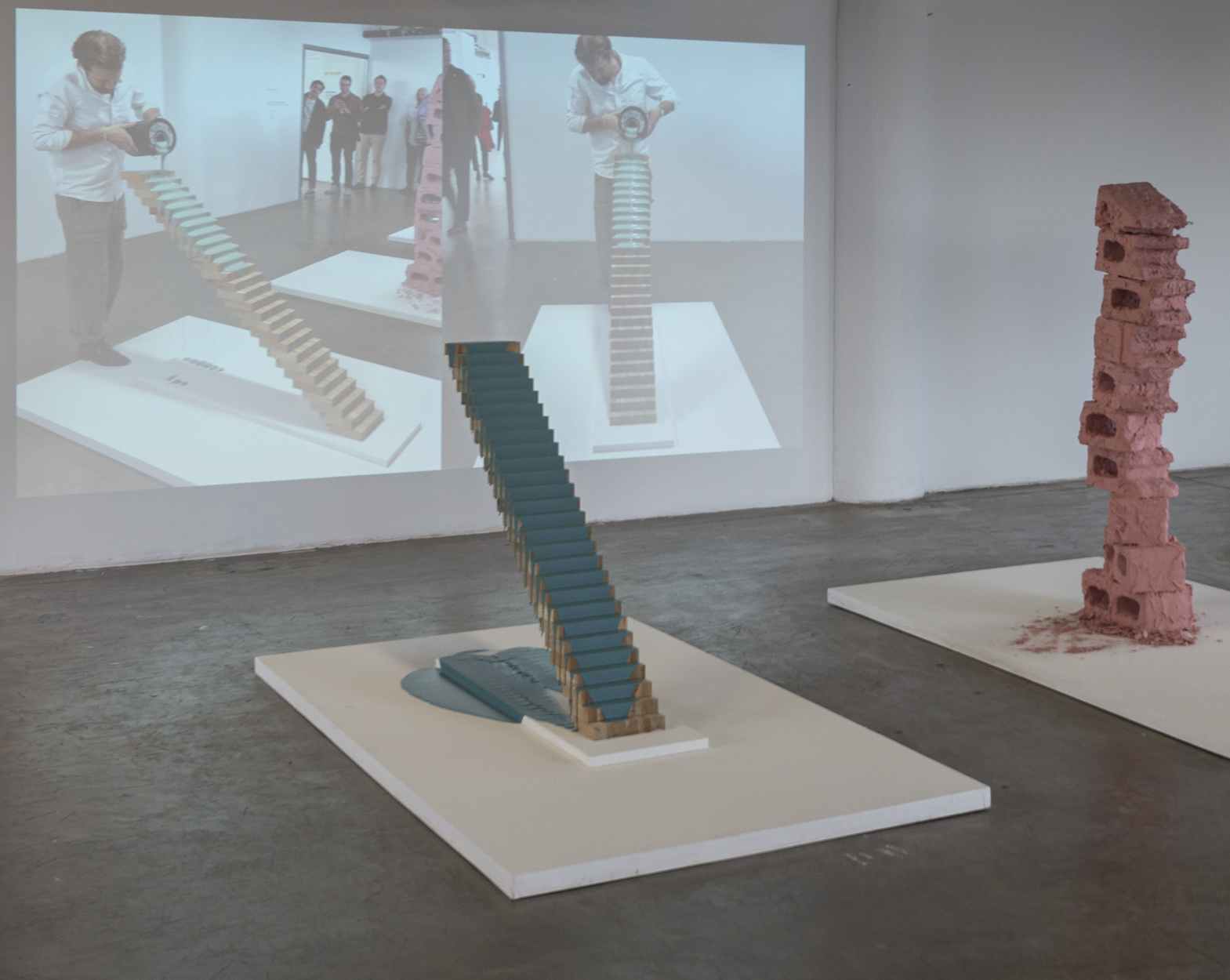 The image shows an art exhibition space with two sculptures on white display platforms. On the left, there is a staircase made of stacked wooden pieces and painted in teal. On the right, a pink sculpture resembling a stacked brick structure is displayed. In the background, a projection depicts a person constructing a similar teal staircase in an art context, alongside a group of spectators observing. The floor is concrete, and the walls are white.