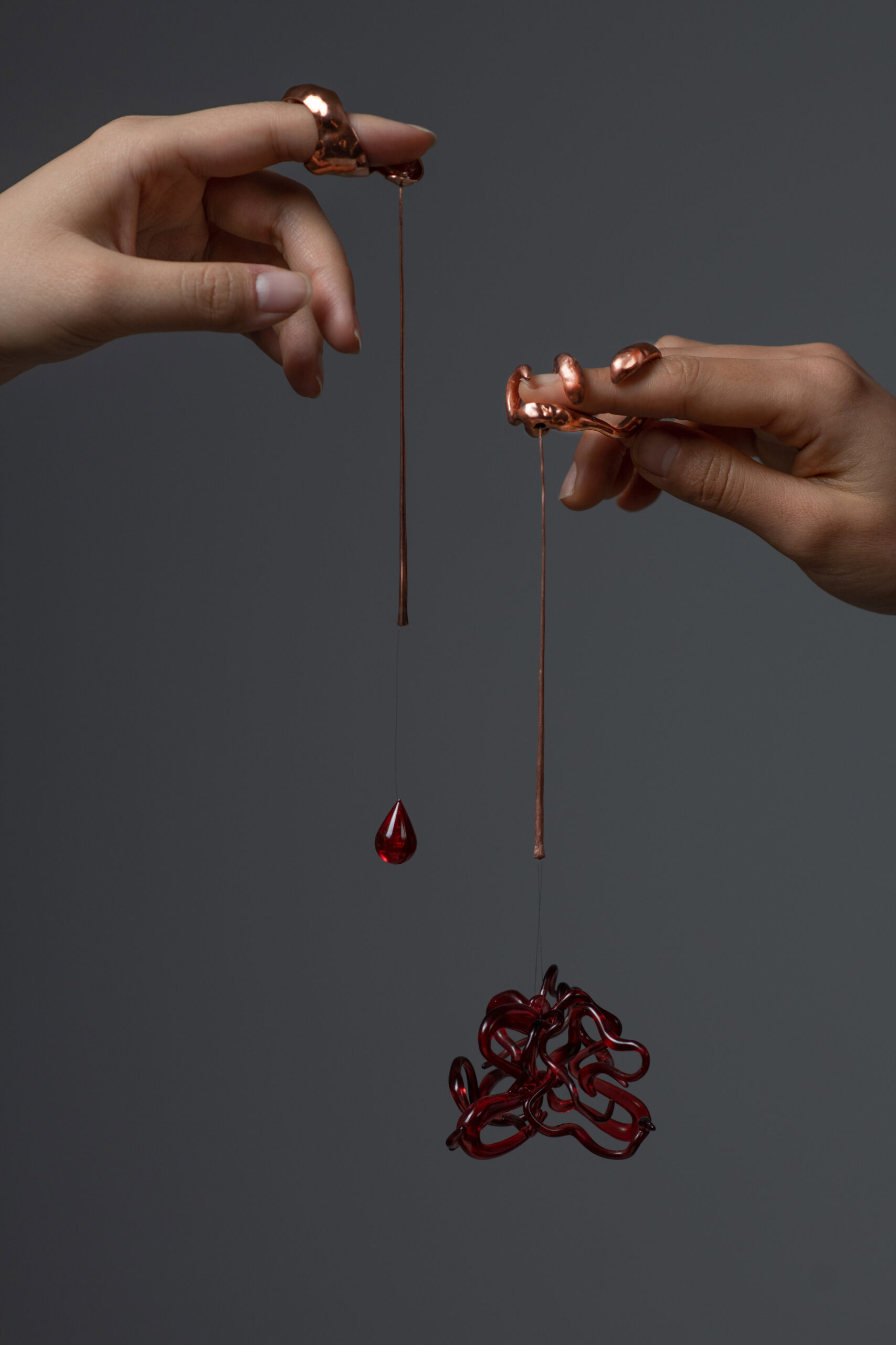 Two hands are holding unique metallic accessories, each shaped like a stylized finger ring. One hand holds a copper string from which a red teardrop hanging ornament is suspended. The other hand holds a tangled red glass-like form, also hanging from a string. The backdrop is a muted gray, emphasizing the intricate details of the accessories and the contrasting textures between the materials.